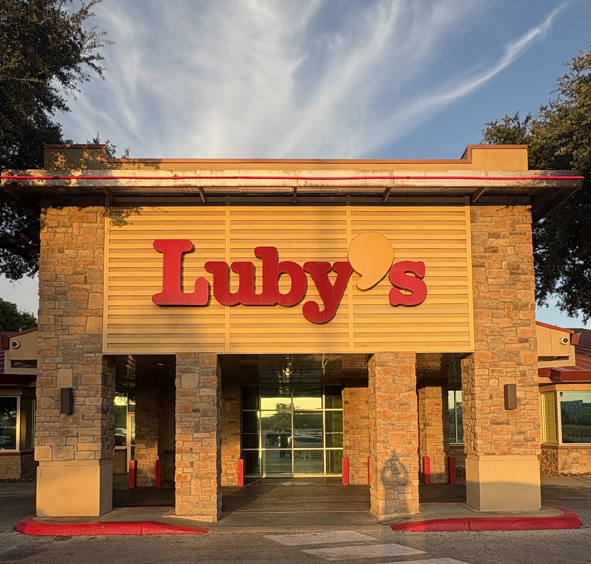 Front entrance of a store with brick facade, large windows, and a sign that reads partially 'Luby's' with a yellow circle and red lettering, during sunset with clear sky and trees surrounding.
