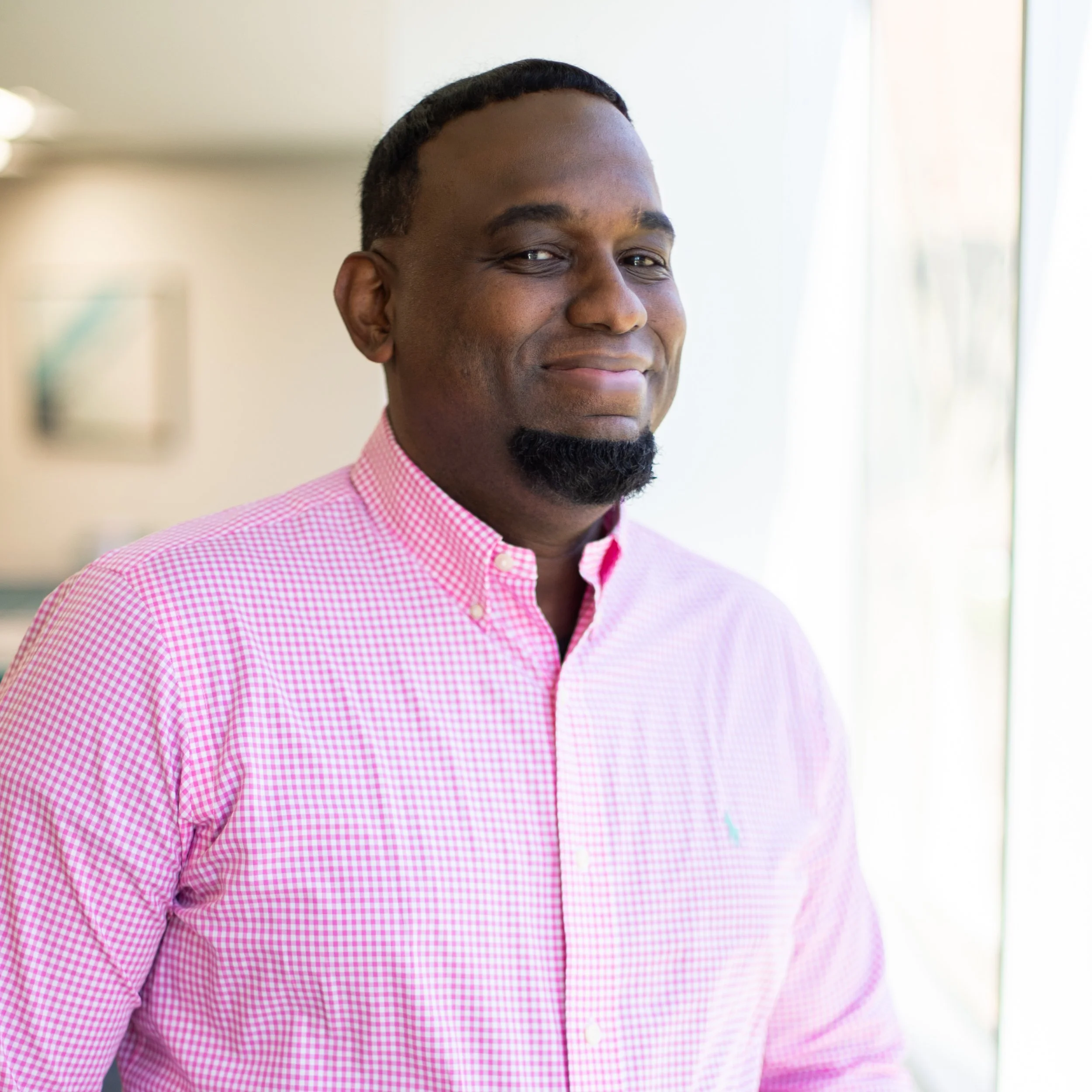 Close-up of a smiling African American man wearing a pink checkered dress shirt, standing indoors near a window with natural light.