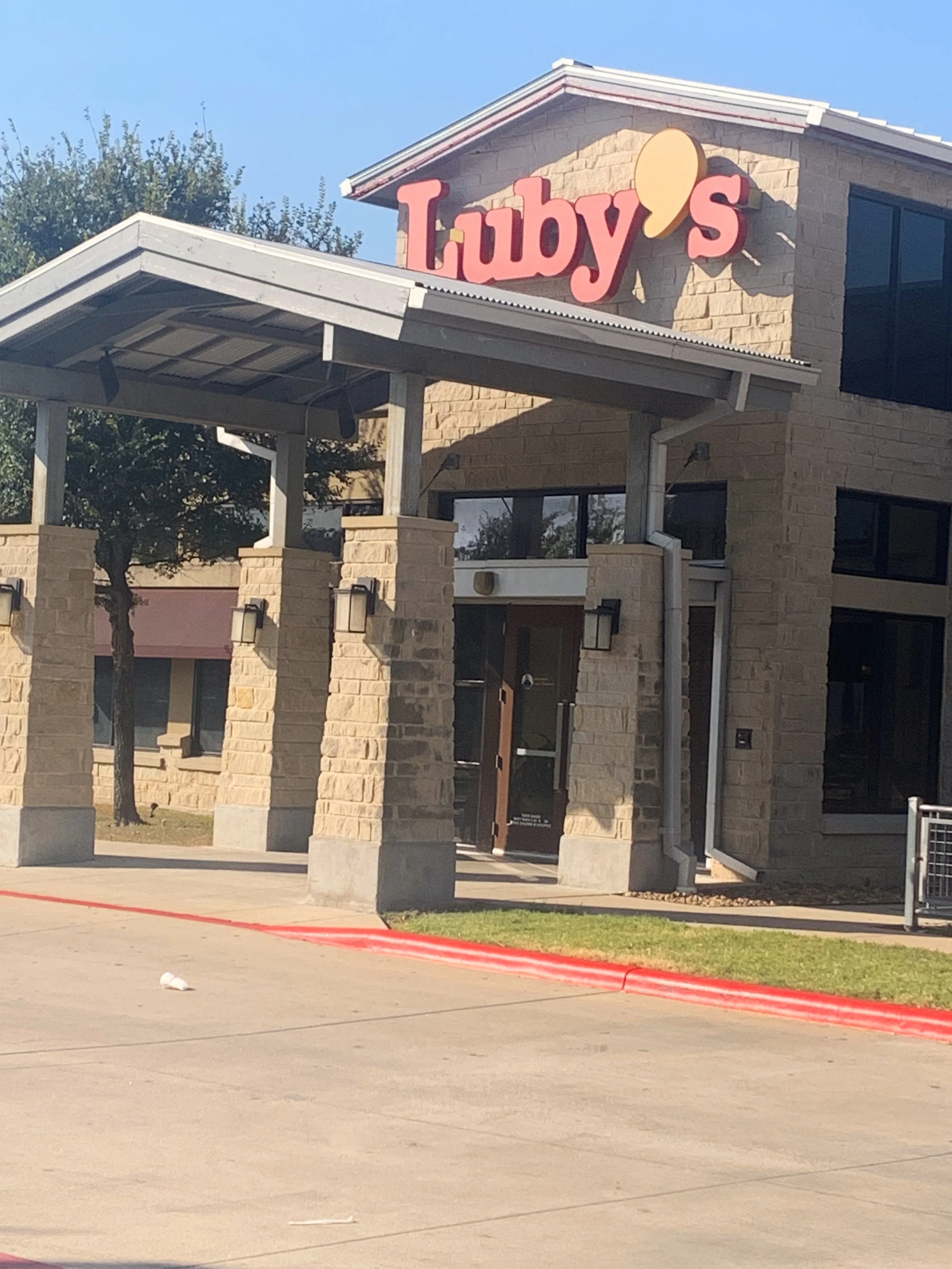 A Starbucks coffee shop with stone walls, a covered entrance, and a sign that says 'Luby's' with a speech bubble shape, on a sunny day.