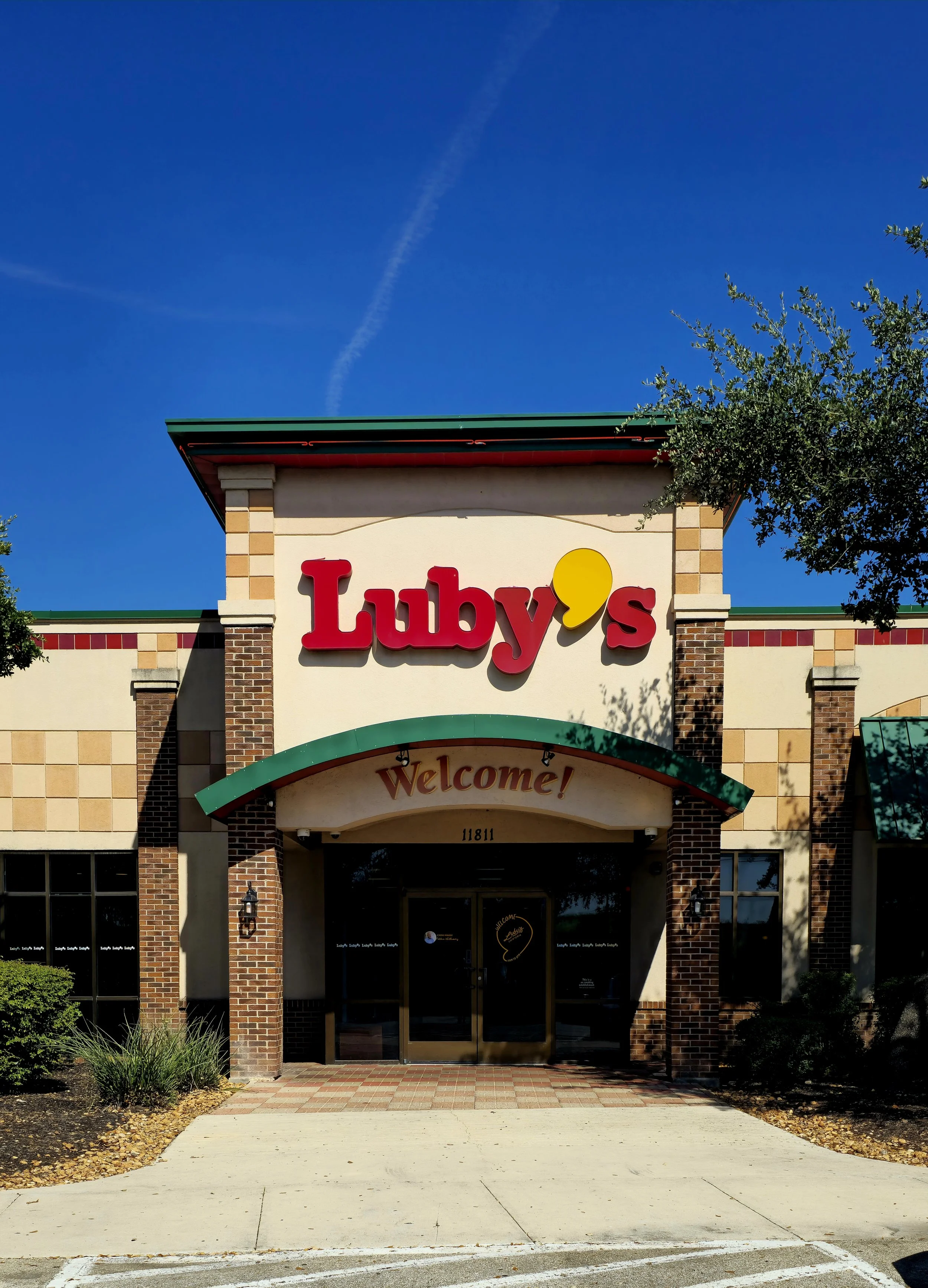 Exterior of a building with a sign that reads 'Luby's' and a welcoming sign below it. The building has brick and beige siding, with a green awning and a paved walkway leading to the entrance. There are trees and small plants around the entrance, and the sky is blue with some clouds.