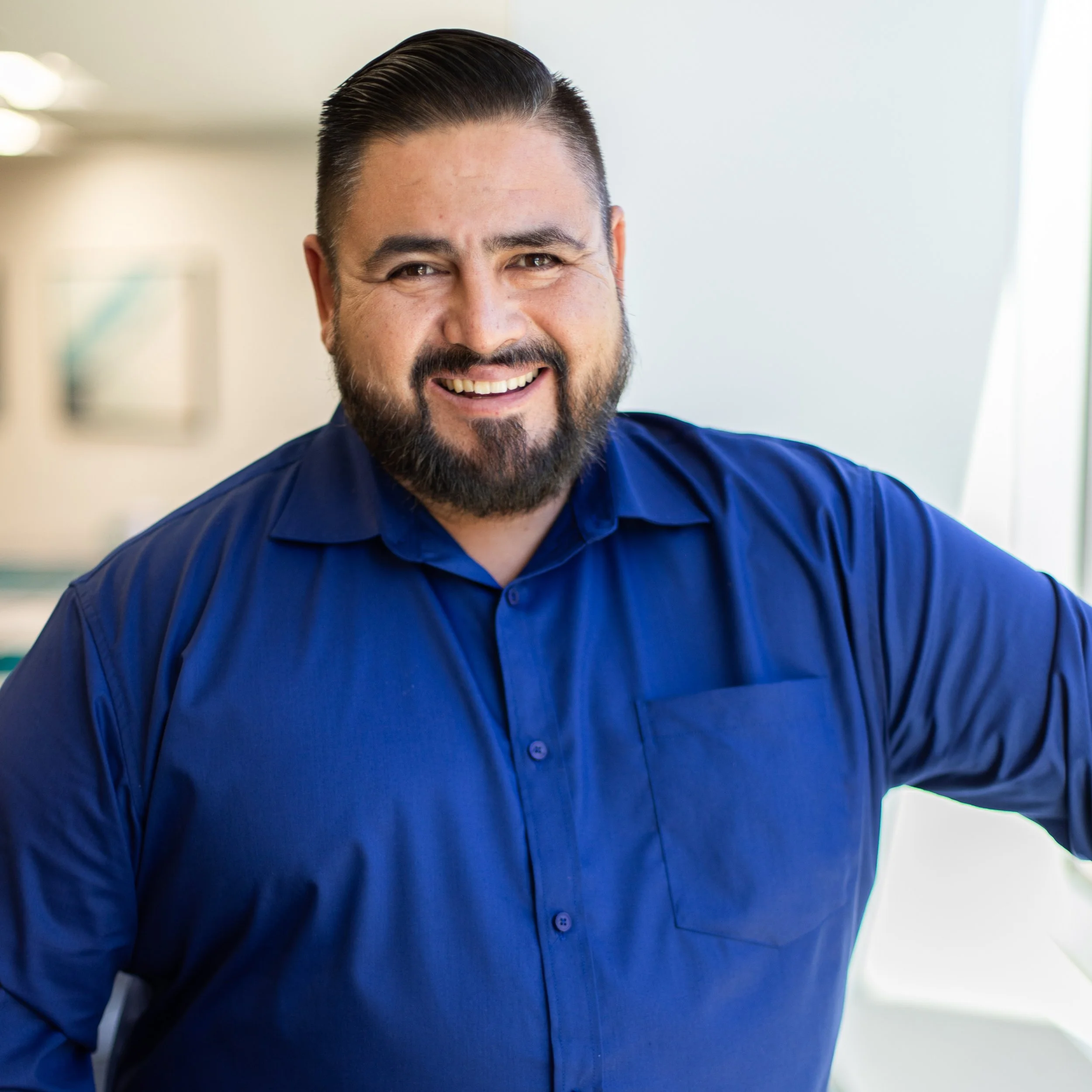 A man smiling in professional attire, wearing a dark blue shirt, in a modern office setting.