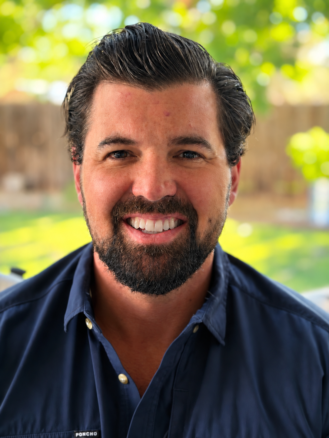 Close-up of a smiling man with dark hair and beard, wearing a navy blue collared shirt, standing outdoors with blurred trees and greenery in the background.