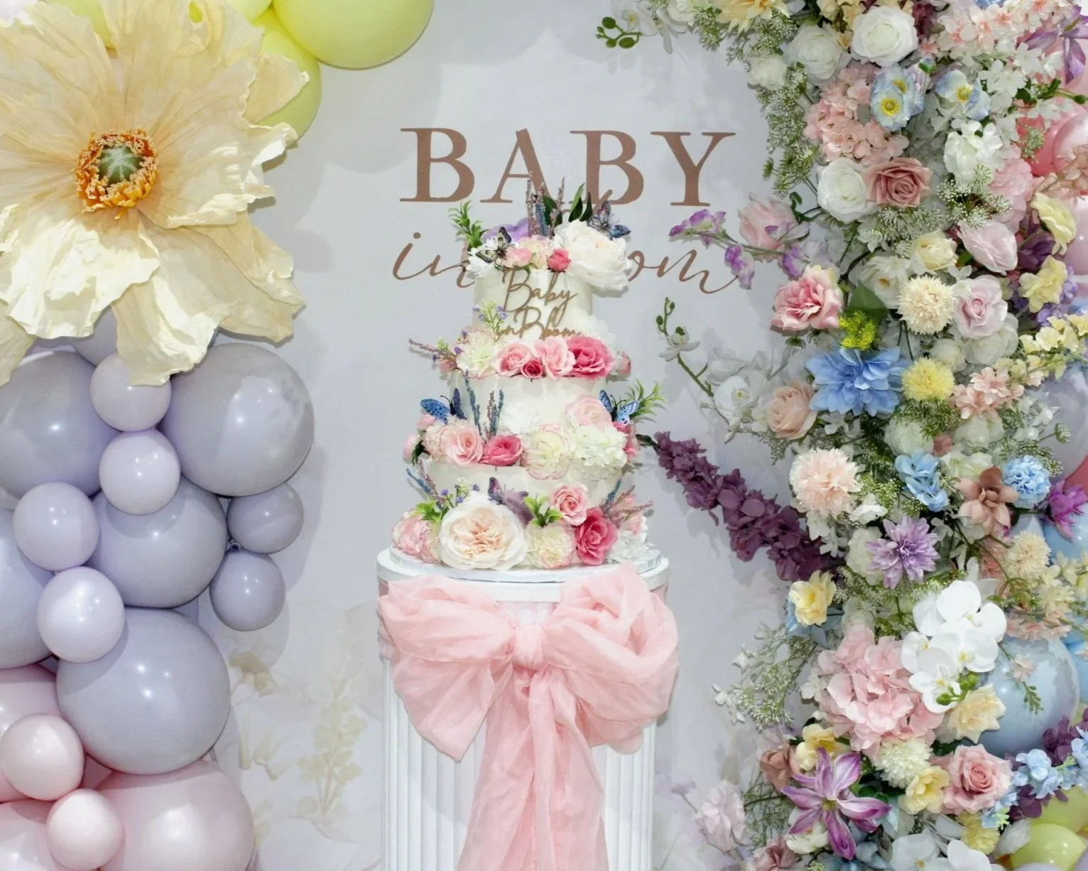 A decorative setup for a baby shower with a floral cake, balloon arrangement, and a large flower wall with pastel-colored flowers, and a sign that reads 'Baby in Bloom'.