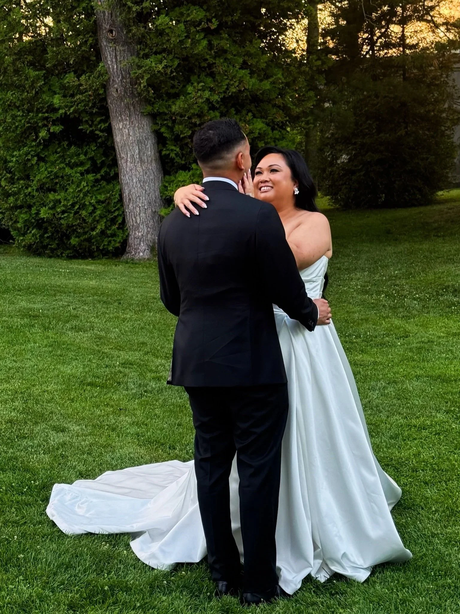 A bride and groom embrace in an outdoor wedding photoshoot on a grassy area with trees in the background during sunset.