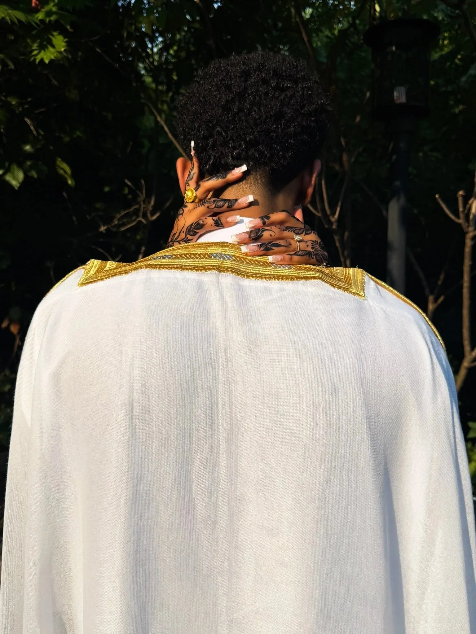 Person dressed in white with gold trim on the shoulder, seen from behind, outdoors with green foliage in the background.