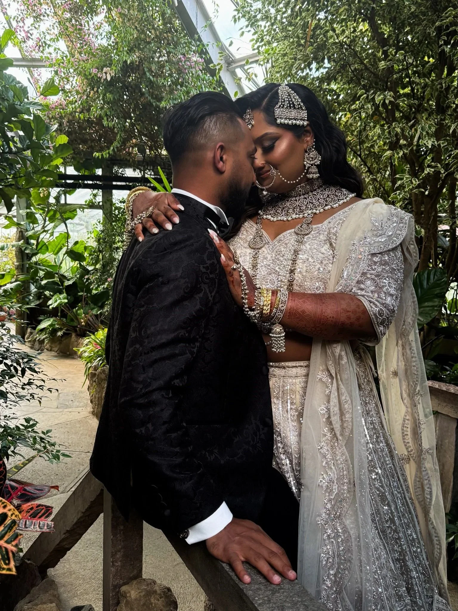 A couple dressed in traditional Indian wedding attire sharing a close, intimate moment in a lush, green garden.