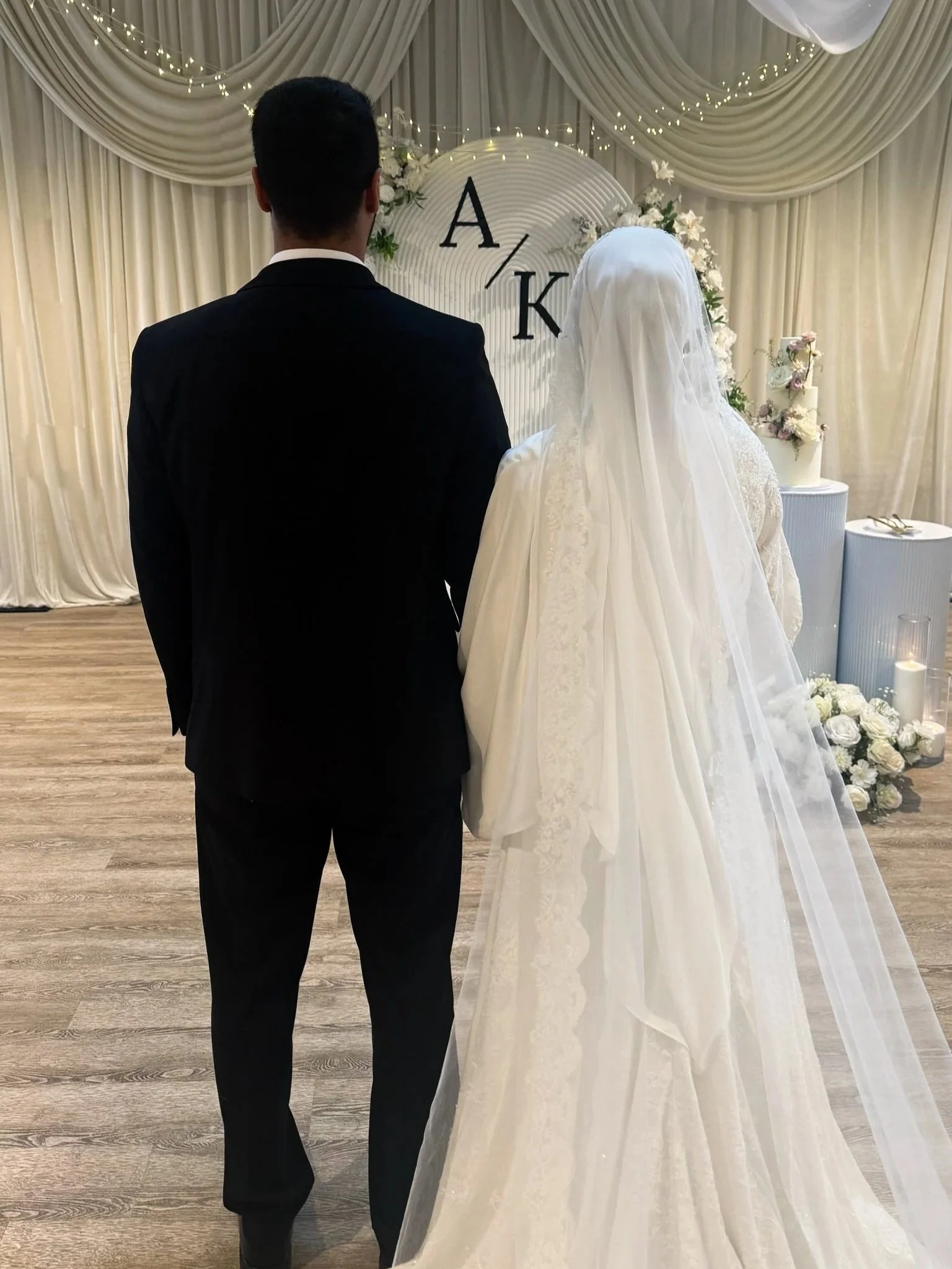 A bride and groom facing away, standing in front of a decorated backdrop with draped curtains, fairy lights, flowers, and a cake, during a wedding ceremony.