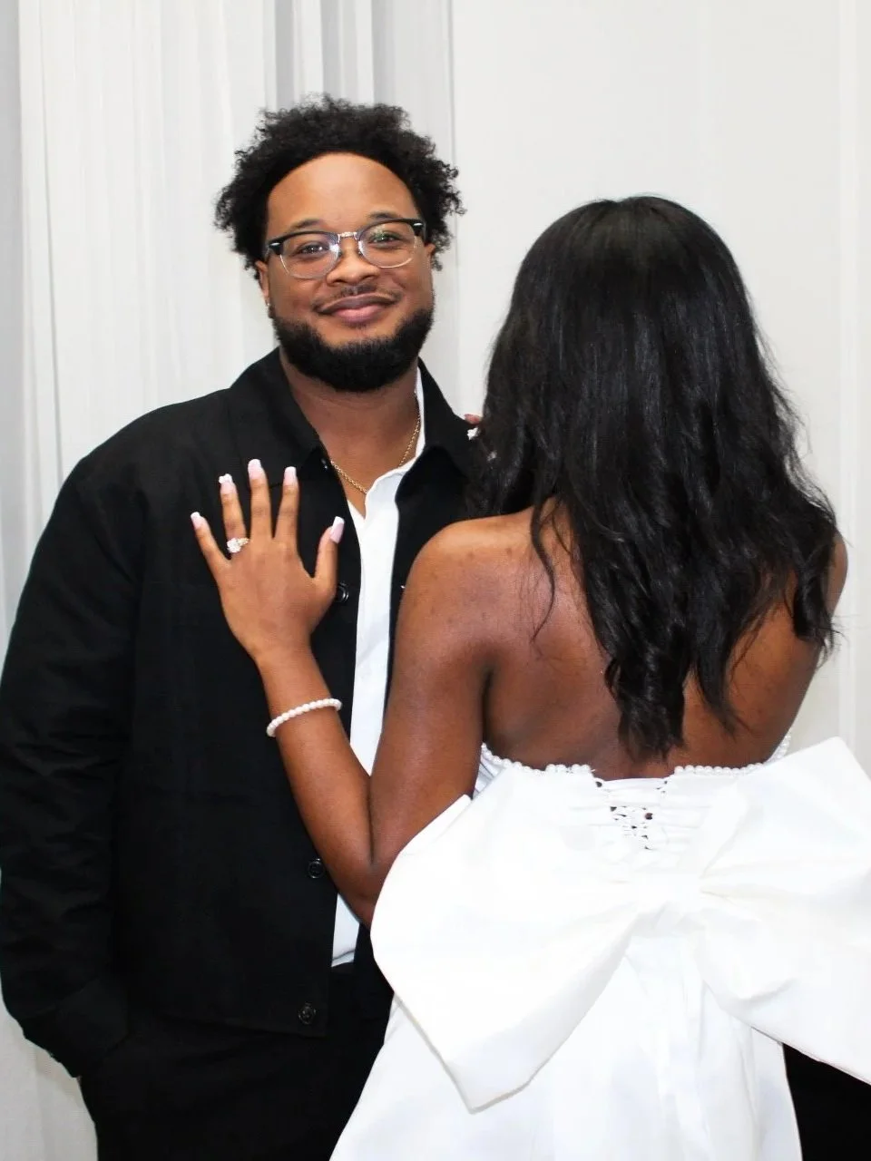 A man with glasses and a beard dressed in a black shirt, standing and smiling, as a woman with long curly black hair in a white strapless dress, facing away, has her hand on his shoulder.