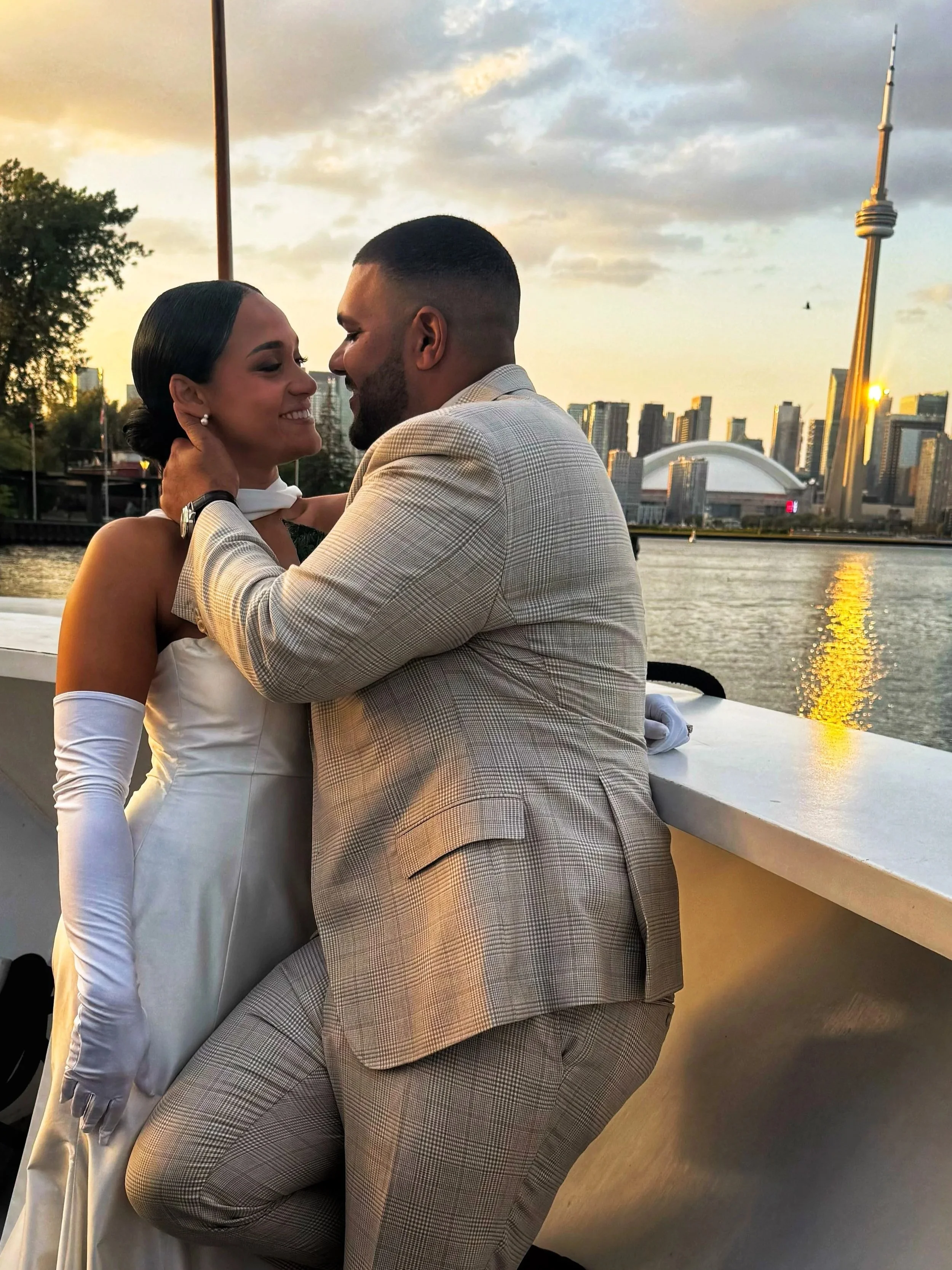A couple sharing a kiss on a boat with the Toronto skyline, including the CN Tower, in the background during sunset.