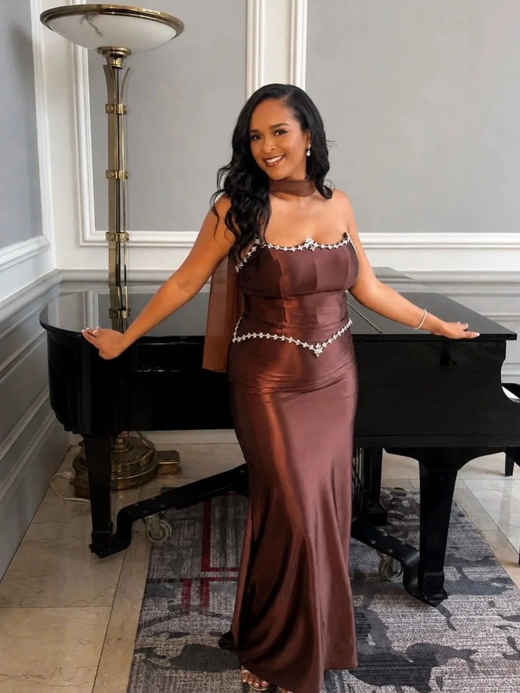 A woman wearing a brown satin evening gown with floral embellishments, standing next to a black grand piano in a room with elegant molding and a marble floor.