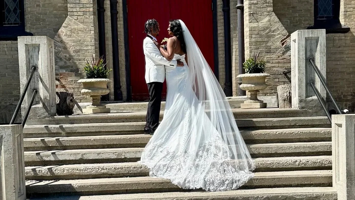 A couple dressed in wedding attire standing on the steps of a brick building, facing each other and holding hands, with the bride in a white wedding gown with a long train and veil, and the groom in a white tuxedo jacket and black trousers.
