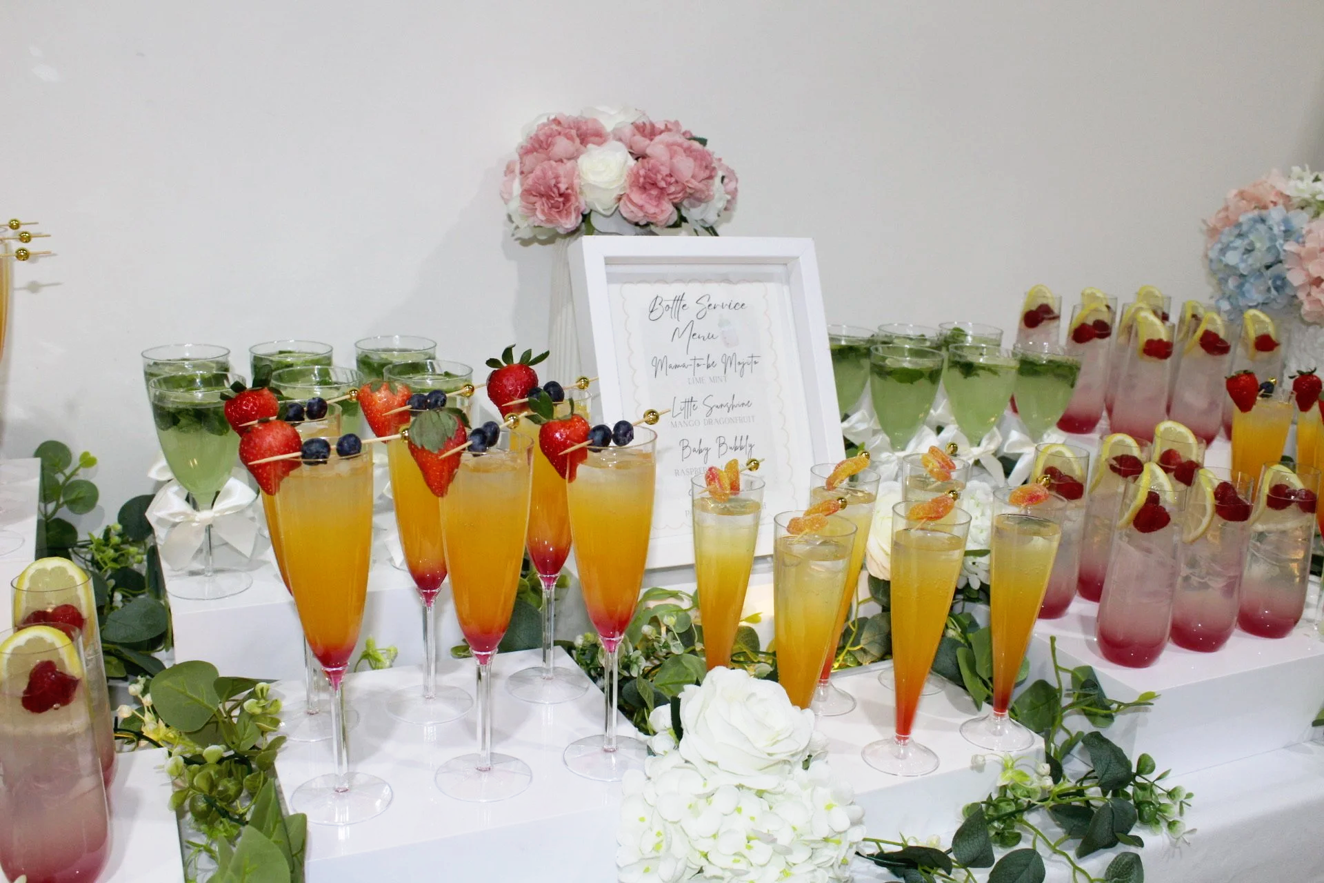 A beverage station with colorful drinks decorated with fruit, surrounded by white flowers and greenery, with a menu sign displayed.