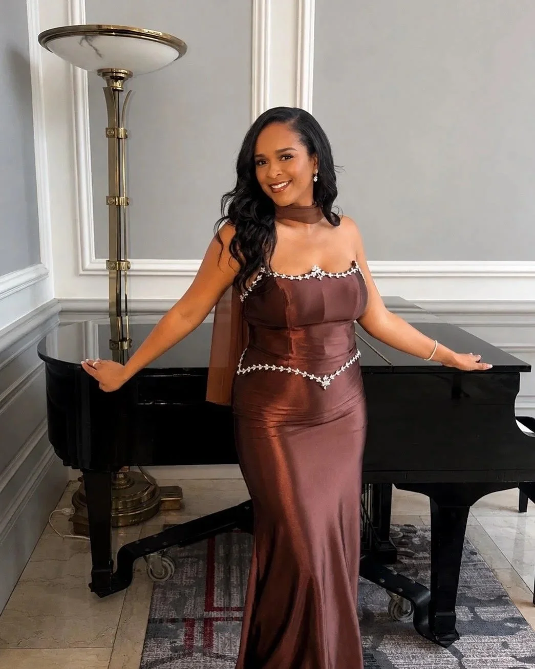 A woman in a brown satin gown with white floral accents stands next to a black grand piano in a room with gray walls and white trim, posing and smiling.