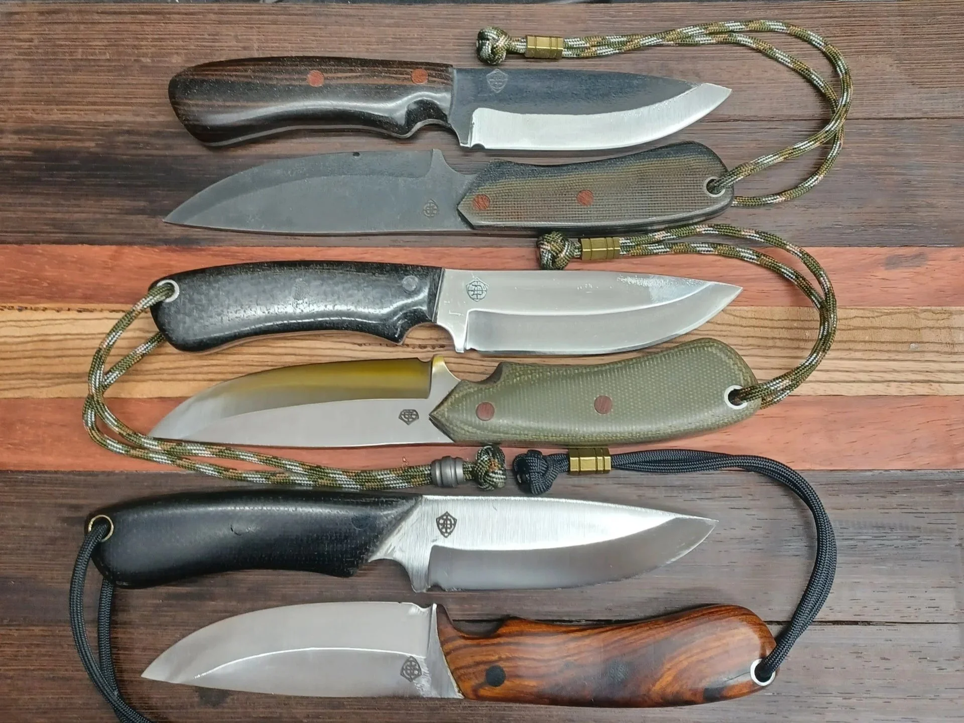 Six hunting knives with various handle materials and lanyards arranged on a wooden surface.