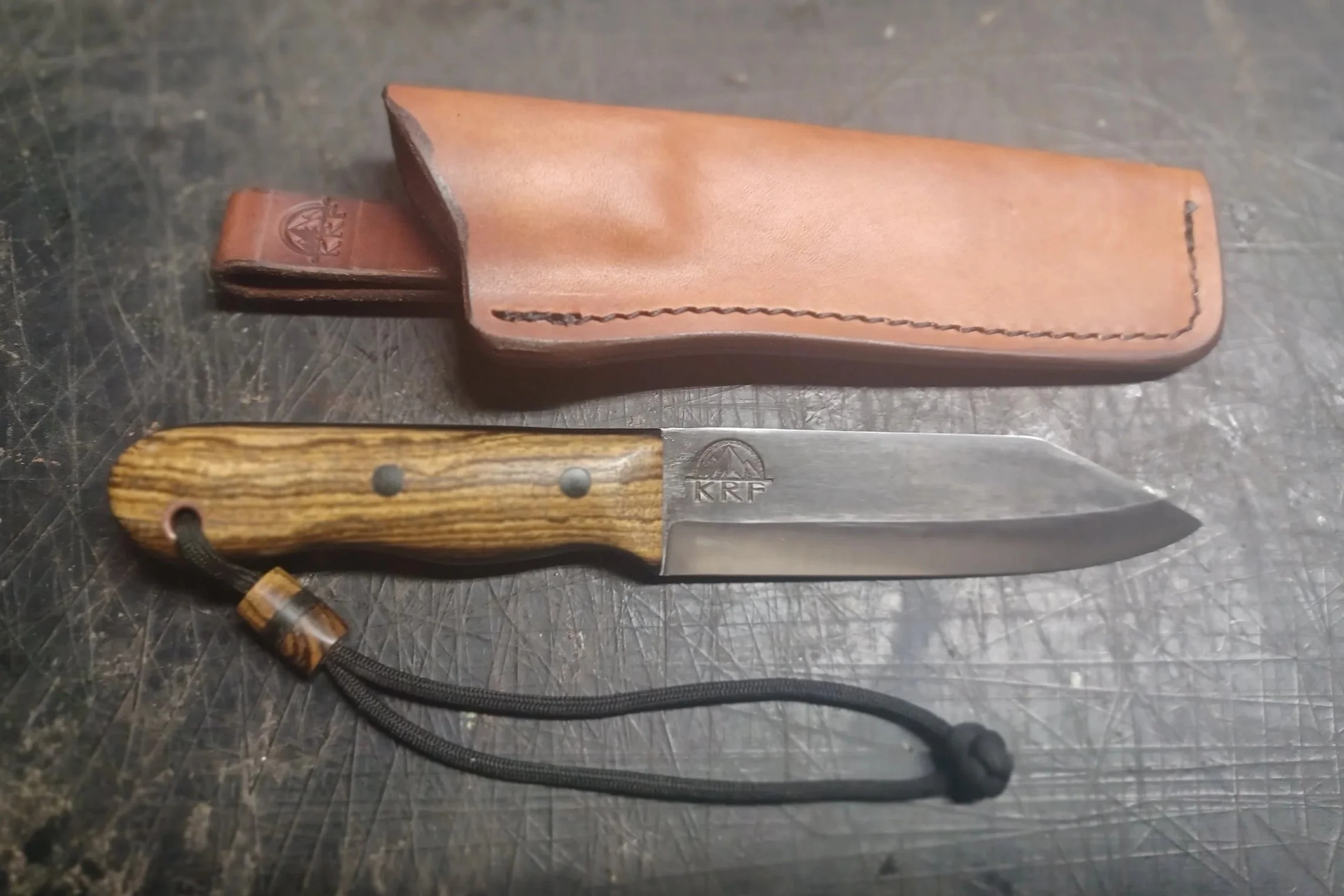 A fixed-blade knife with a wooden handle and lanyard, placed next to a brown leather sheath on a dark surface.