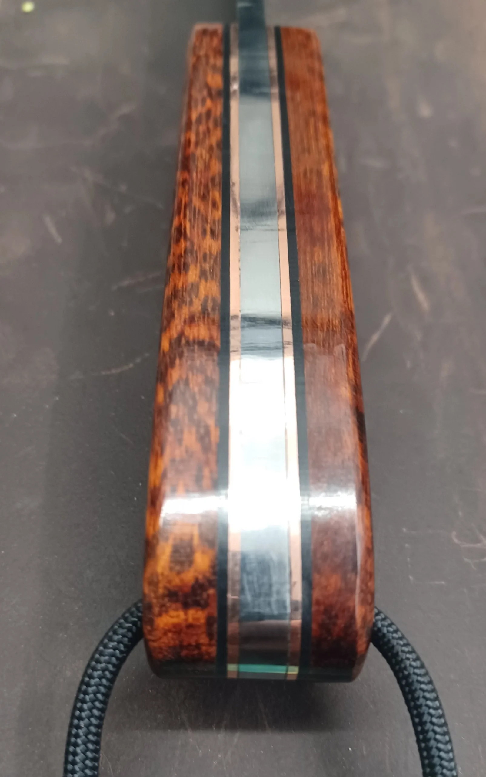 Close-up of a wooden and metal knife handle resting on a black surface, showing the shiny metallic center and the wooden sides with a textured pattern.