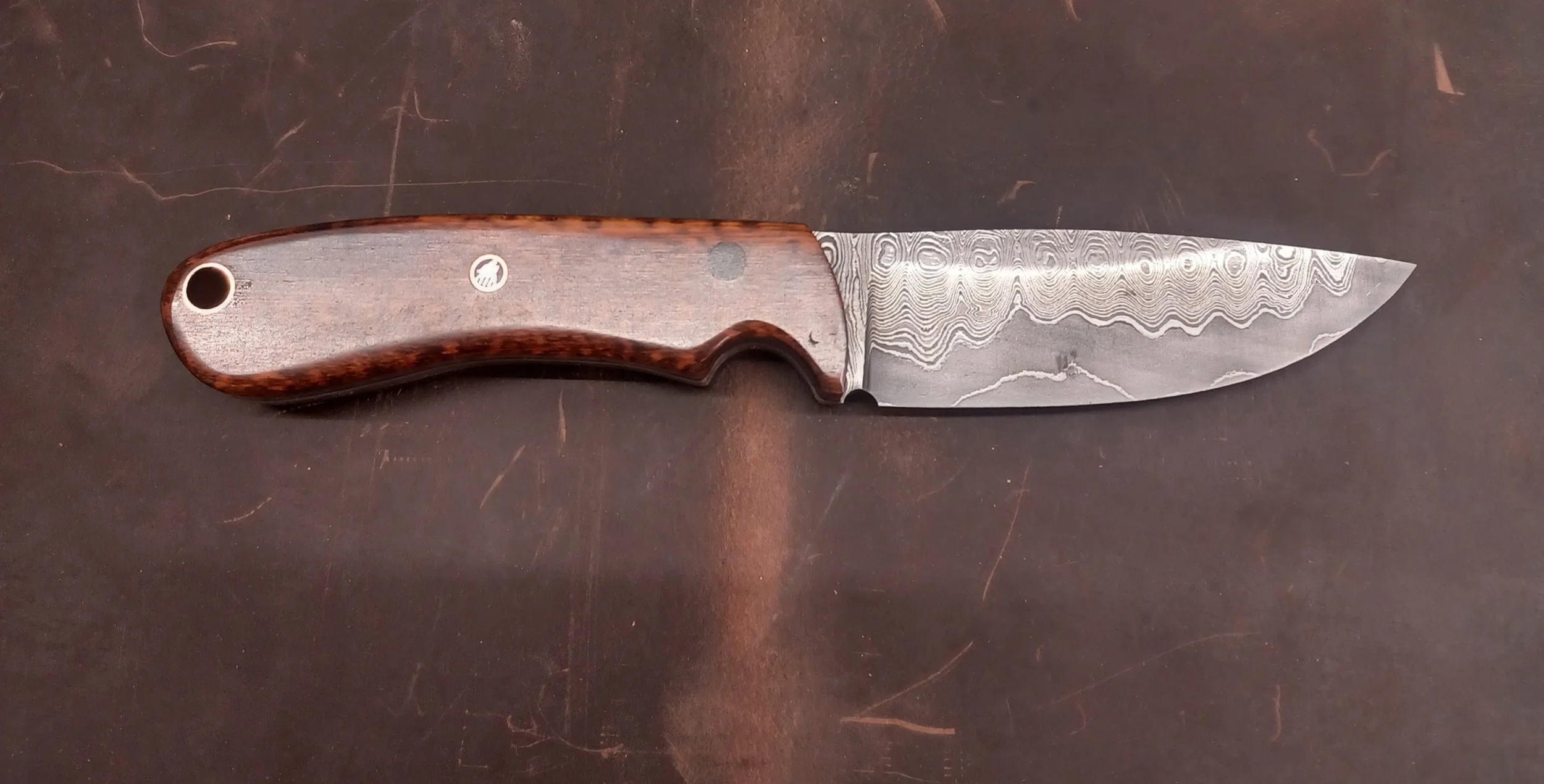 A close-up view of a Damascus steel kitchen knife with a wooden handle, resting on a dark surface.