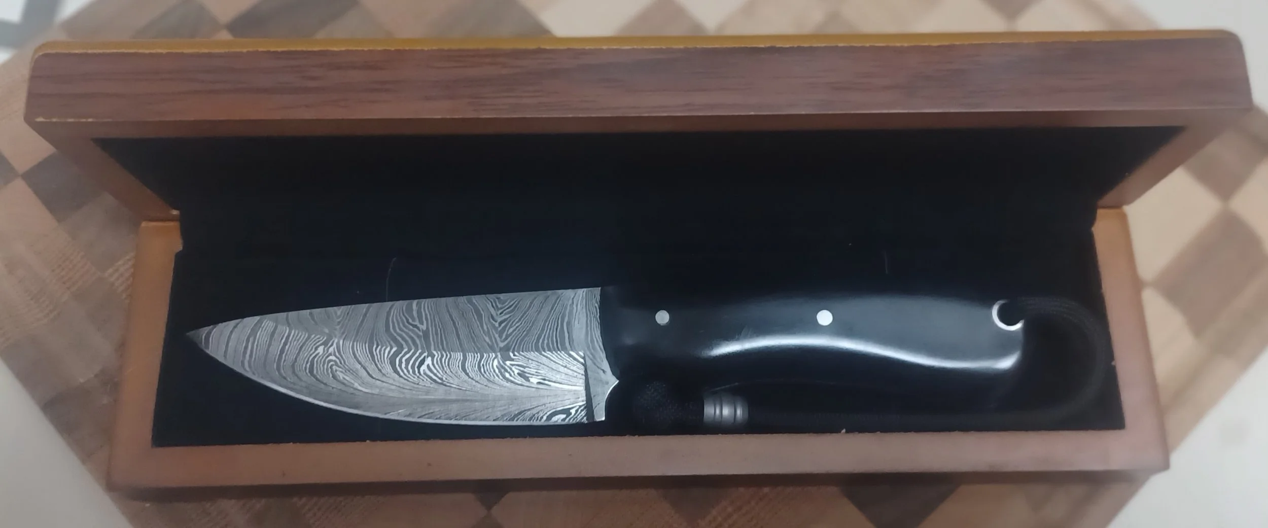 A knife with a patterned blade and a black handle inside an open wooden box with a black interior, resting on a tiled surface.