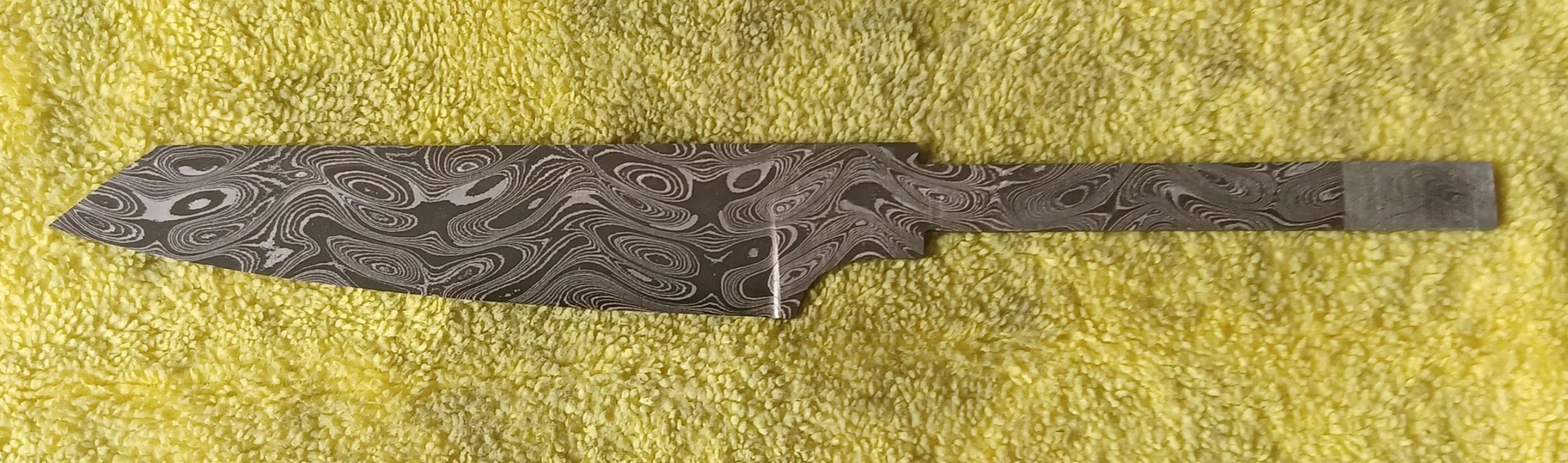 A knife with a marbled, swirling pattern on the blade lying on a yellow textured fabric.