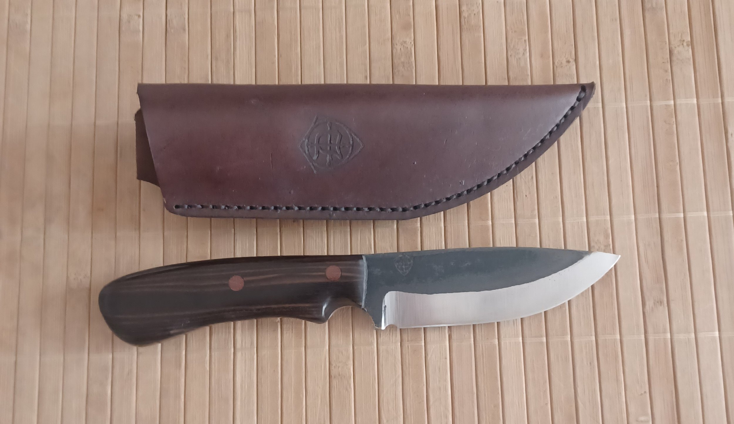 A fixed-blade knife with a dark wooden handle and a leather sheath, placed on a bamboo mat.