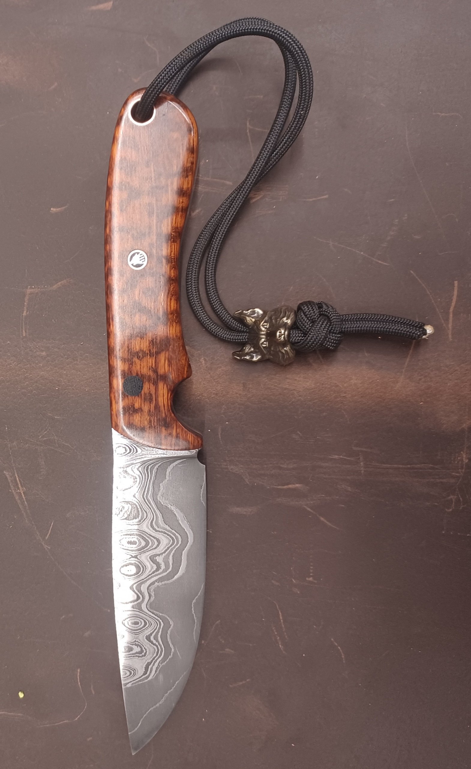A decorative knife with a Damascus steel blade, a wooden handle, and a lanyard with a wolf-shaped metal bead, resting on a dark surface.