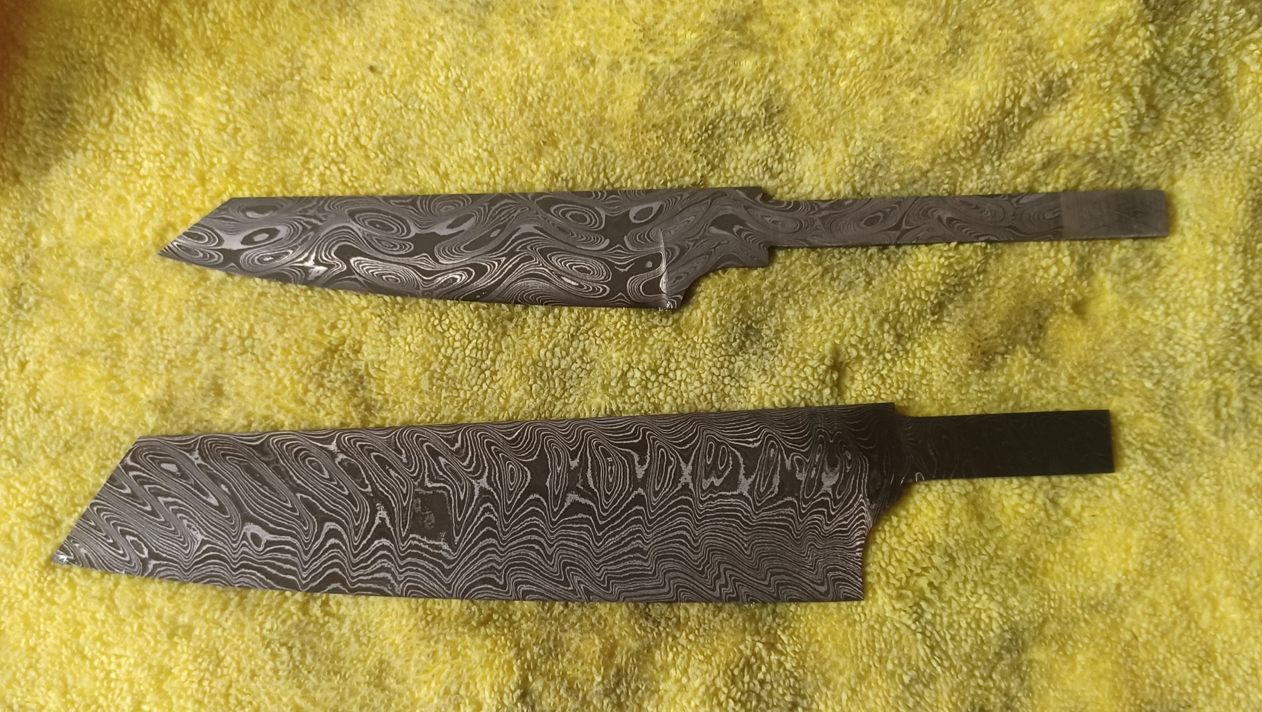 Two Damascus steel knives with intricate swirling patterns on the blades, placed on a yellow textured surface.