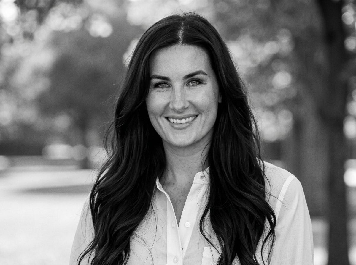 A smiling woman with long dark hair, wearing a light-colored button-up shirt, outdoors with blurred trees in the background.