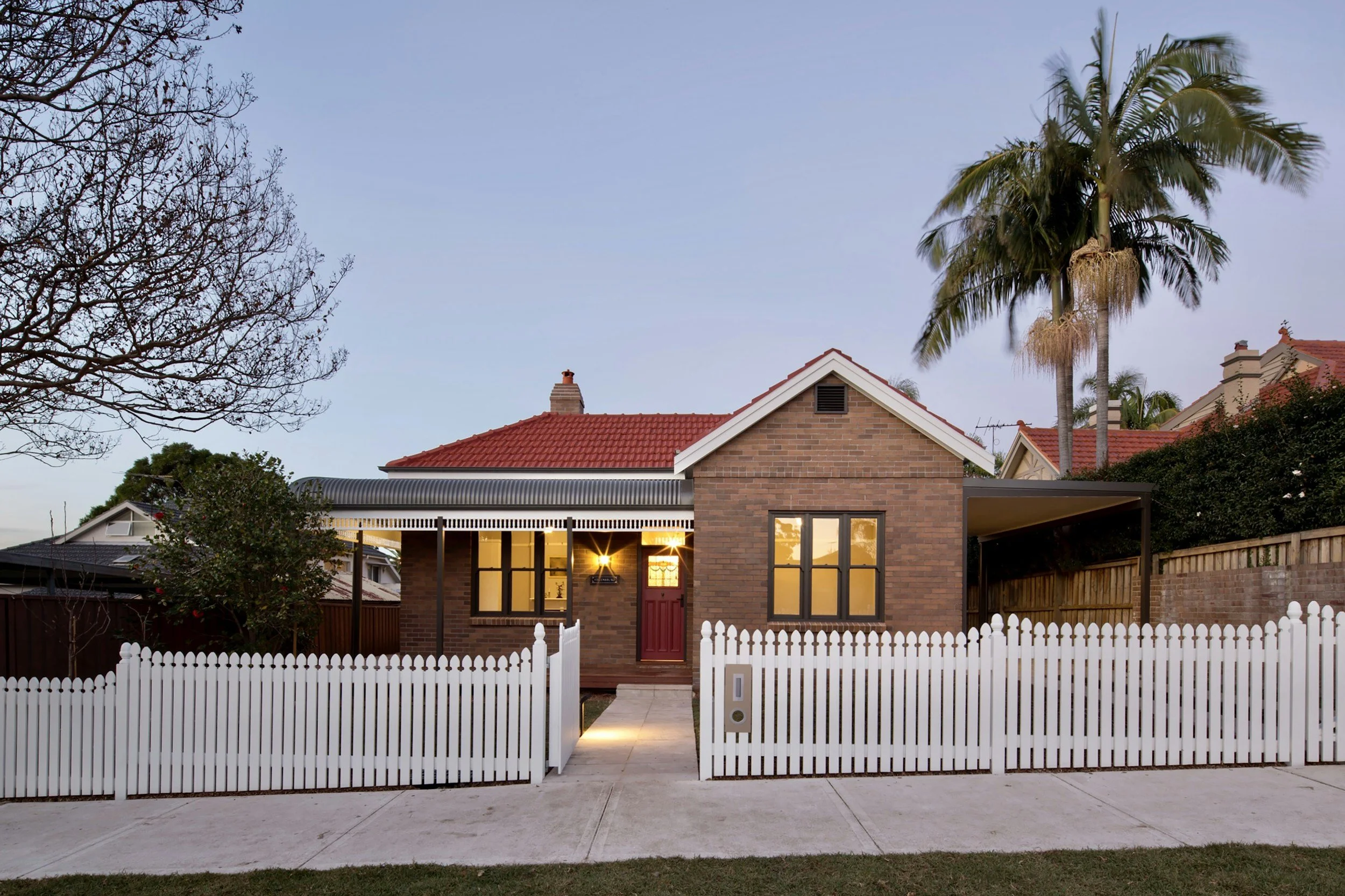 A two-story brick house with a red-tiled roof, large windows, and a white picket fence in front. The house has a small front yard with a concrete walkway leading to the red front door. There are tall palm trees and leafless trees in the background, along with neighboring houses. The sky is clear and the lights inside the house are on.