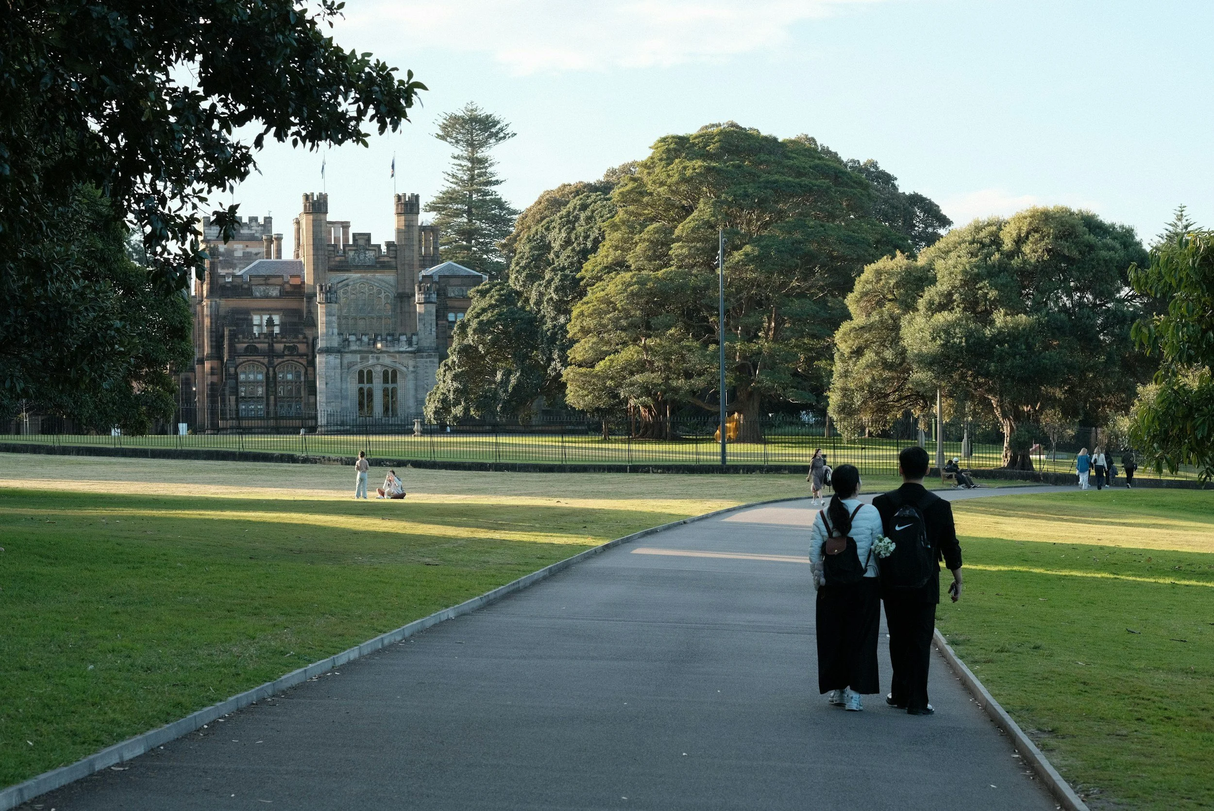 People walking on a paved path through a park with green grass and trees, with a historic castle-like building in the background under a clear sky.
