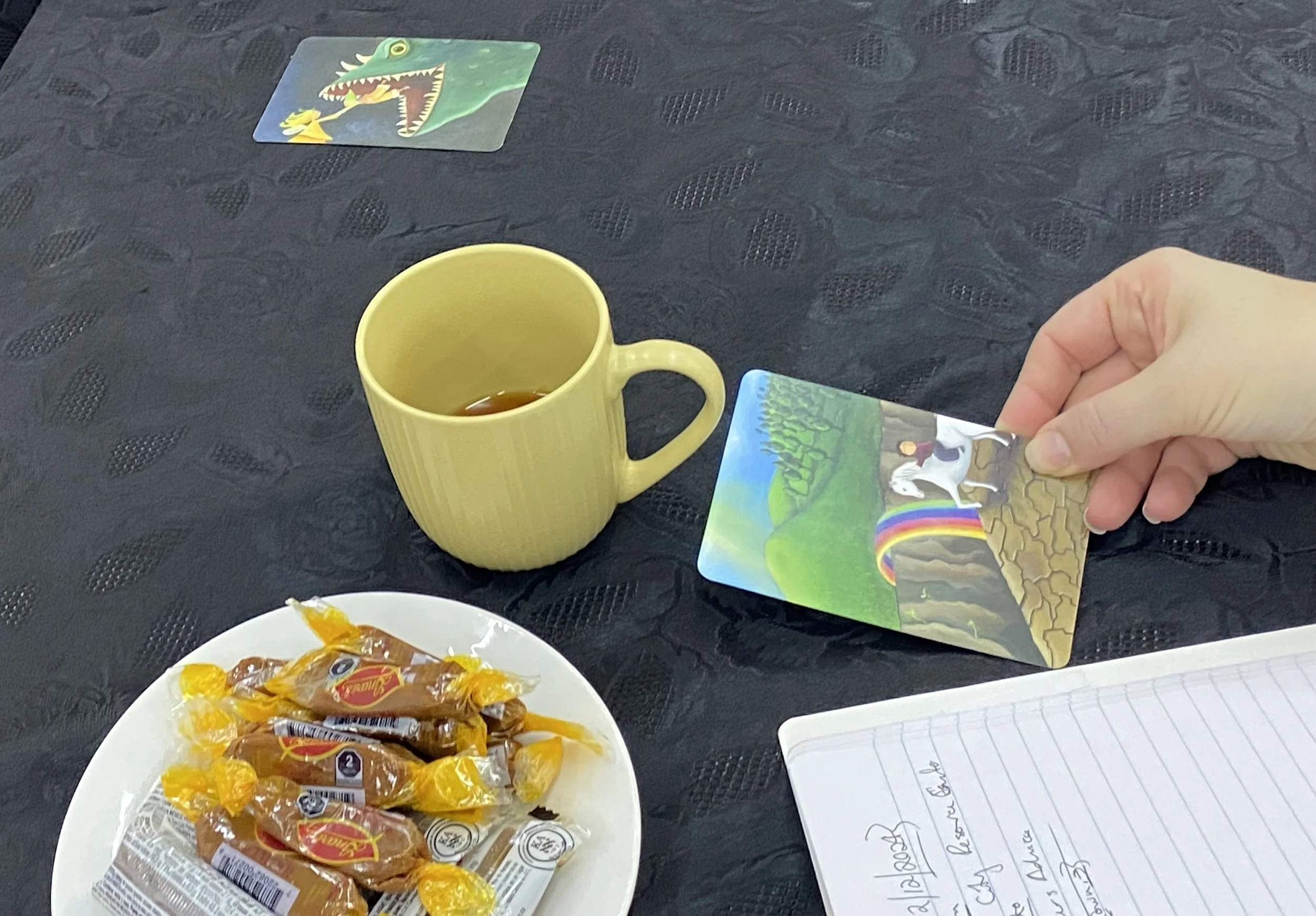 hand holding a dixit card, with a plate of mexican sweets and a coffee cup on the table