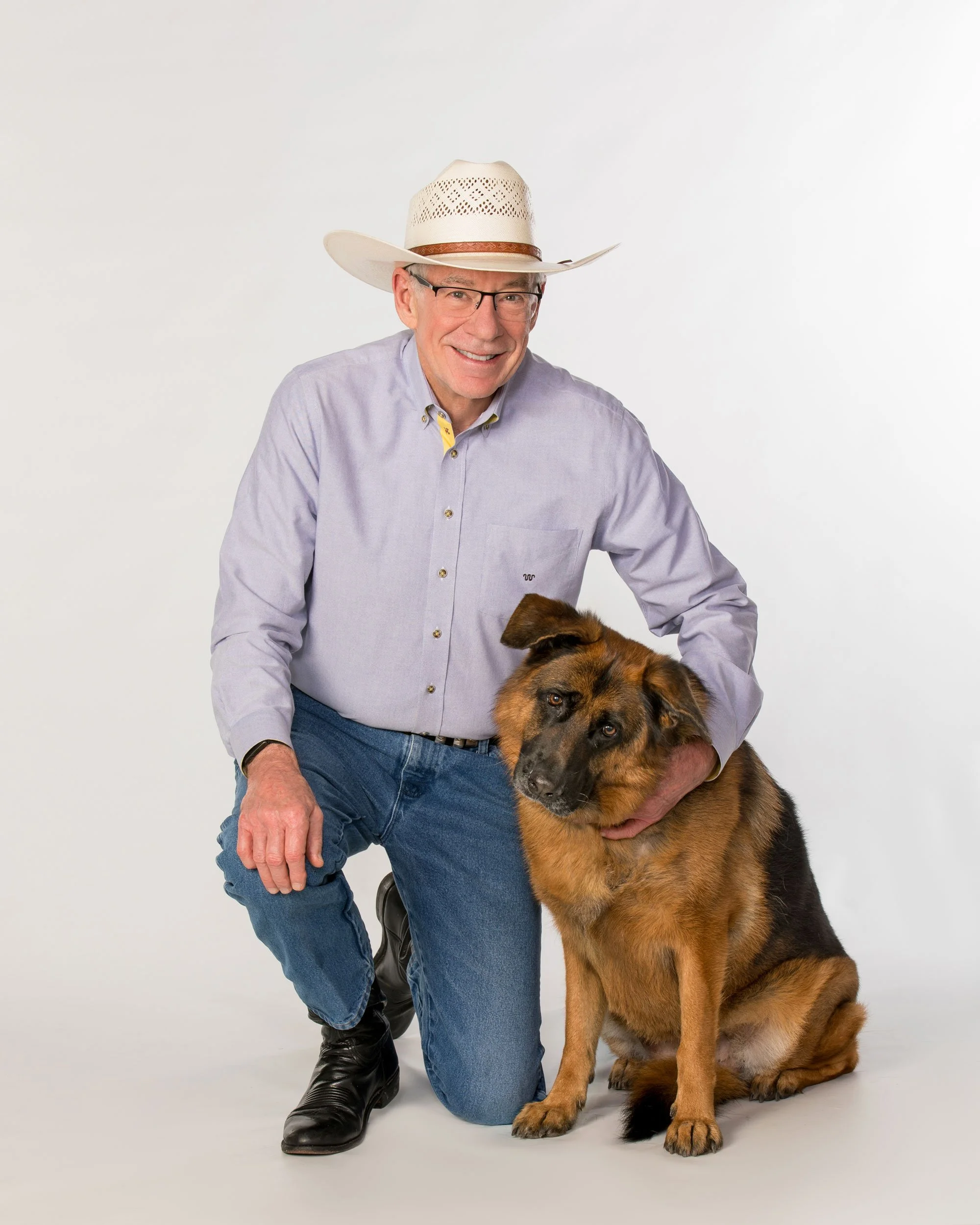 HW Ryan wearing a cowboy hat, glasses, and a light blue shirt kneeling next to a large German Shepherd dog against a plain white background.