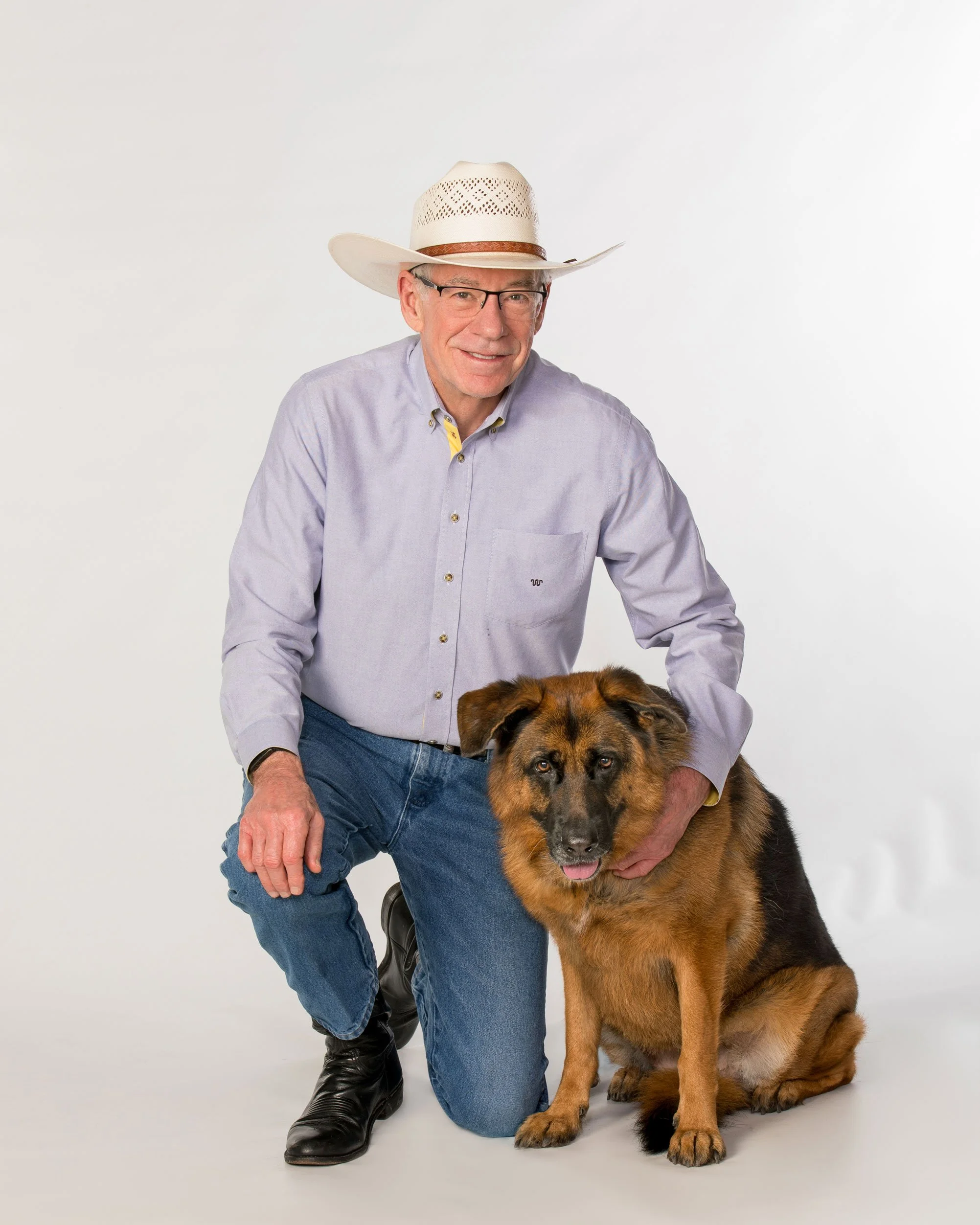 A man wearing glasses, a cowboy hat, and a light purple shirt kneels next to a large German Shepherd dog against a plain white background.