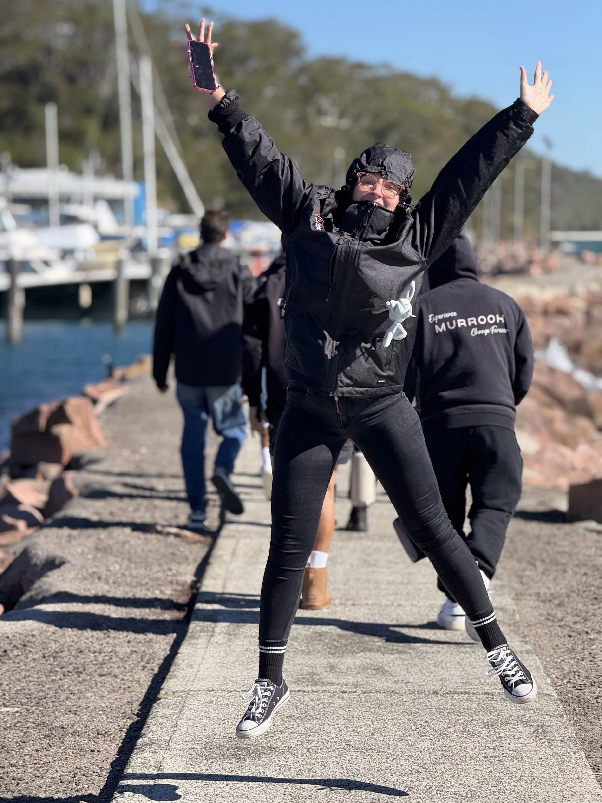 Person jumping with arms raised, wearing black jacket, glasses, and sneakers, at a marina with boats and other people in the background.