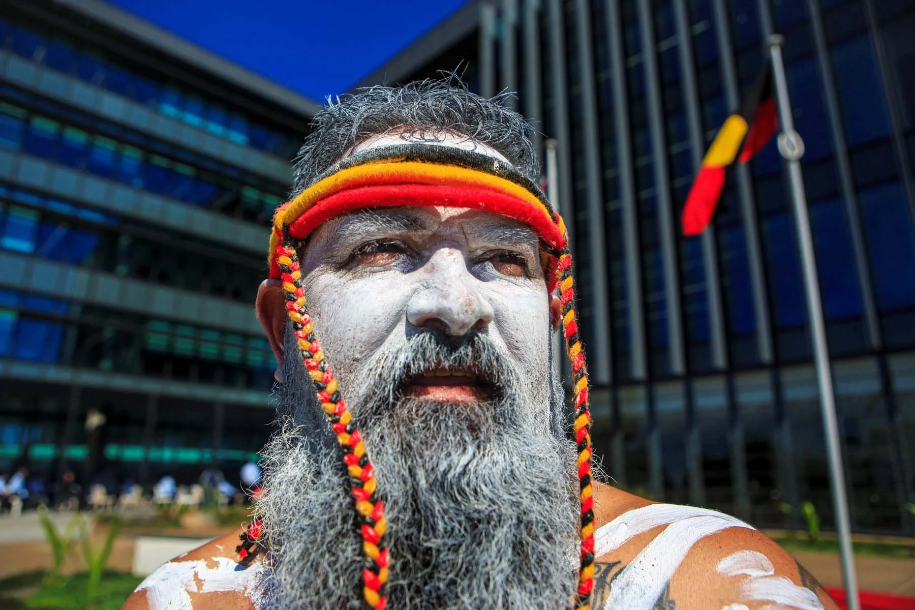 Man with white face paint, wearing a red, yellow, and black headband and traditional attire, standing outdoors with a modern building in the background.