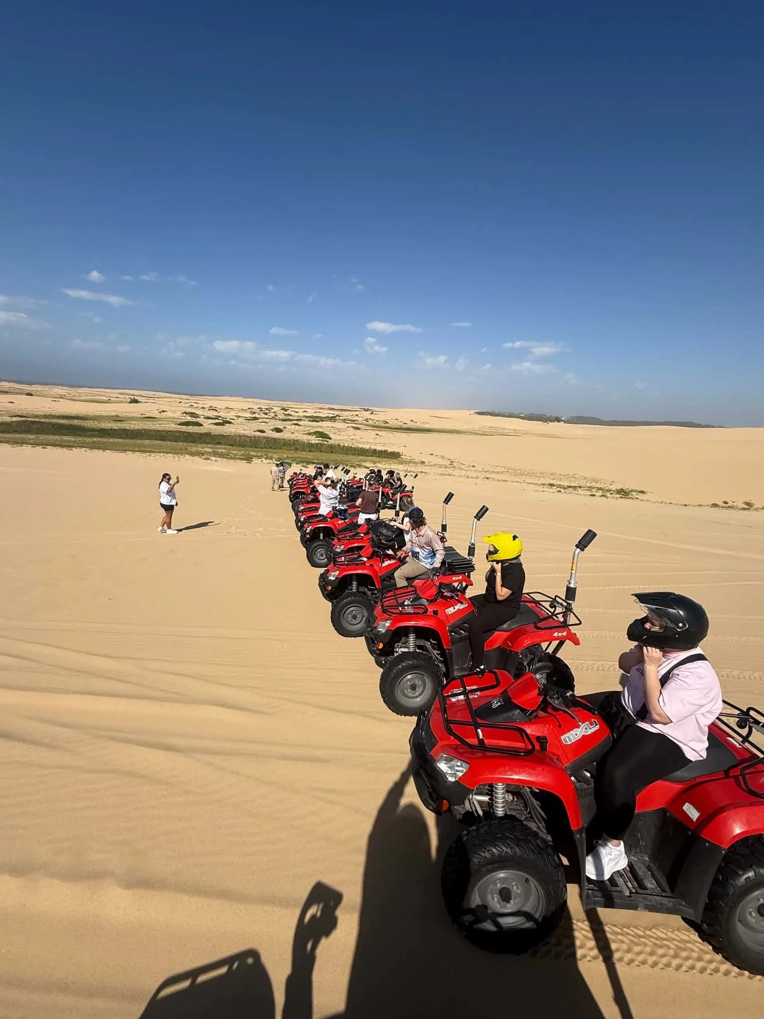 People riding red all-terrain vehicles in a desert with sand dunes under a clear blue sky.