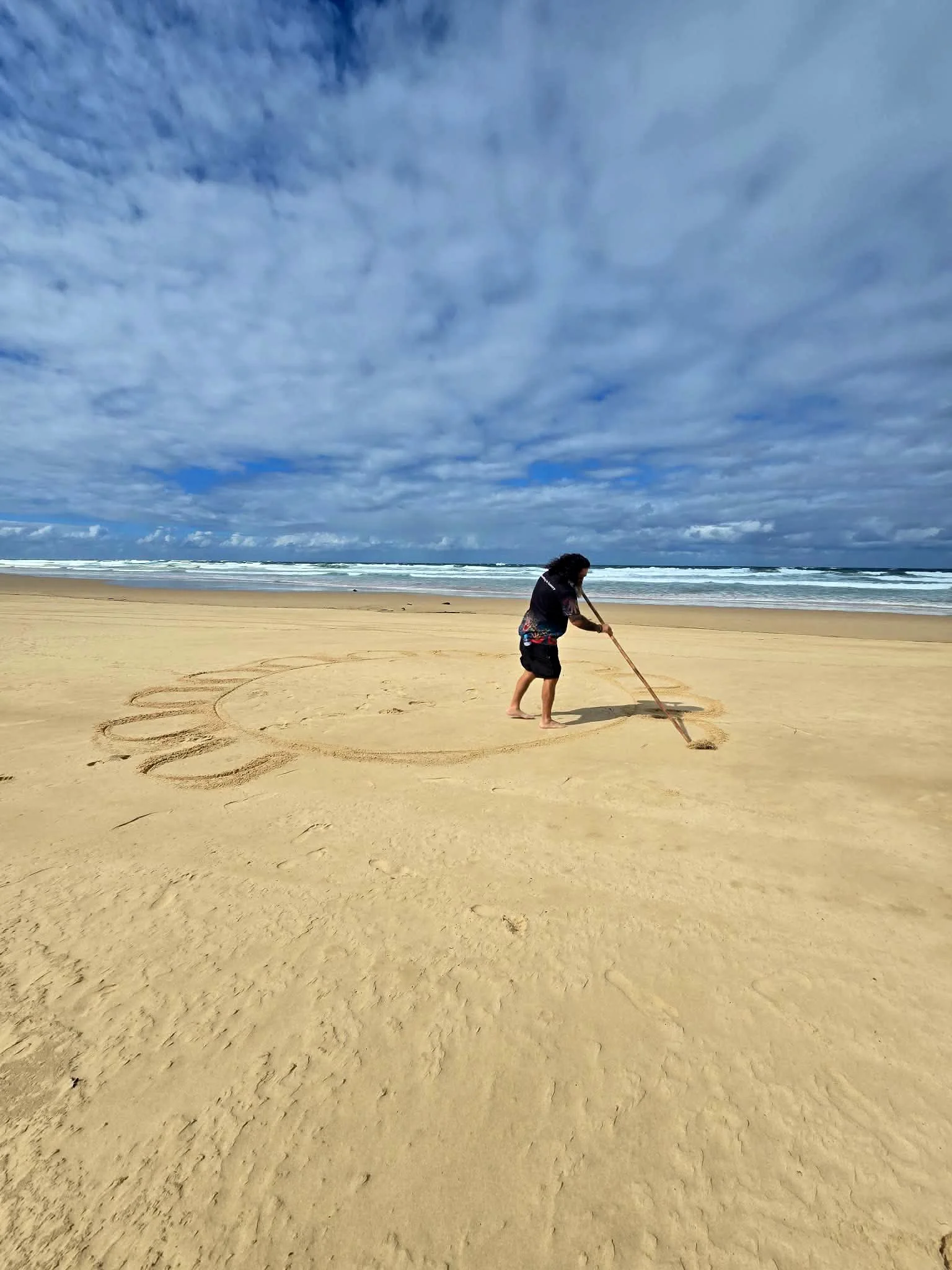 Child playing with a stick on a sandy beach, with ocean waves and cloudy sky in the background.