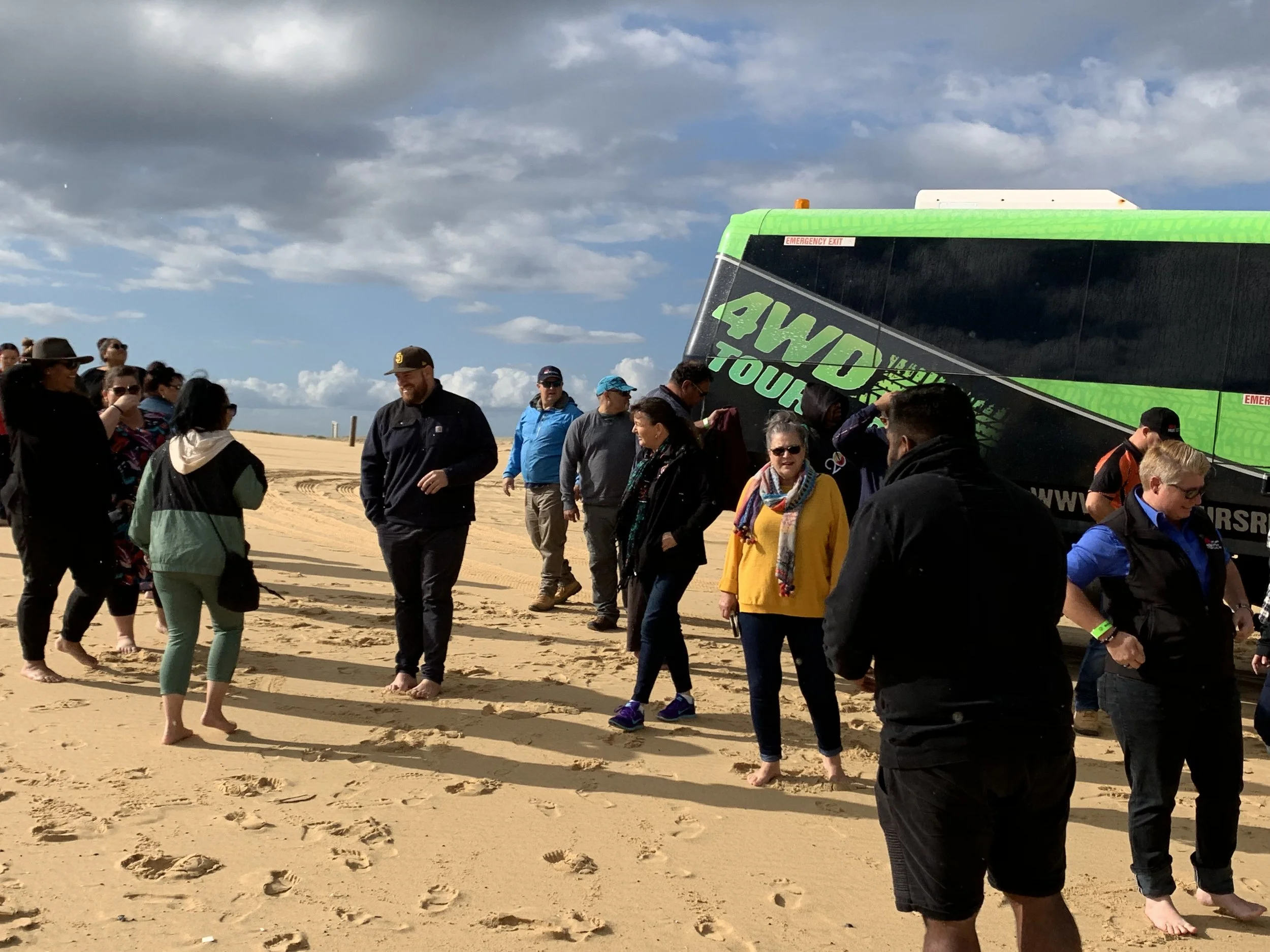 A group of people walking on sandy terrain outdoors, with a large green and black tour bus in the background and a partly cloudy sky overhead.