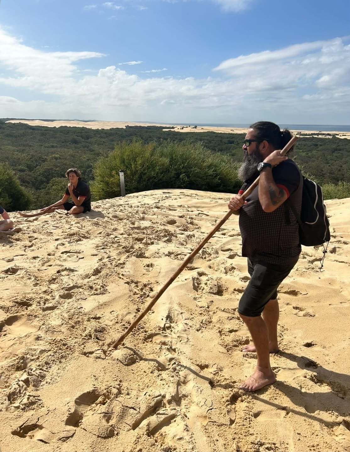 A man with a beard, sunglasses, and a black backpack holds a wooden stick while standing barefoot on sandy terrain in a natural landscape with dunes, green bushes, and a distant ocean under a partly cloudy sky.