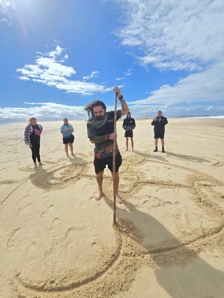 A group of five people on a sandy beach, with one man in the foreground drawing a large circle in the sand with a stick, and the others standing behind him, with a partly cloudy sky above.