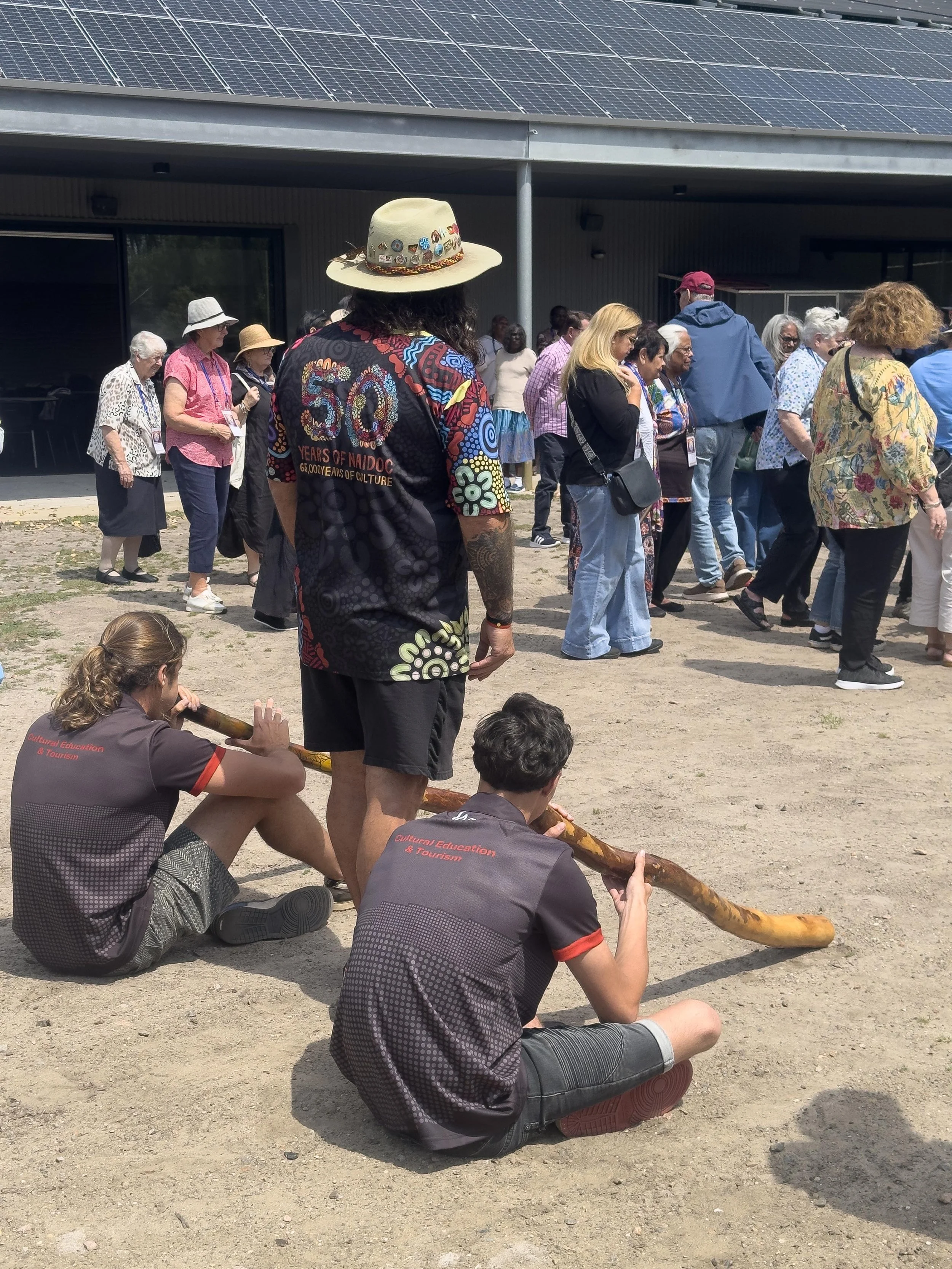 A crowd gathered outside a building and two people seated on the ground playing traditional didgeridoo instruments, with others standing and observing.