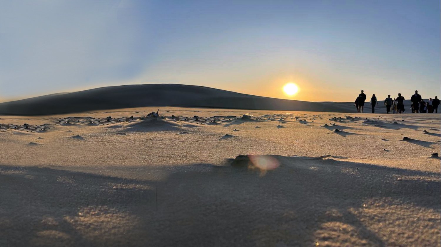 Sunset over a desert landscape with a group of people walking along the horizon, sand dunes in the foreground.