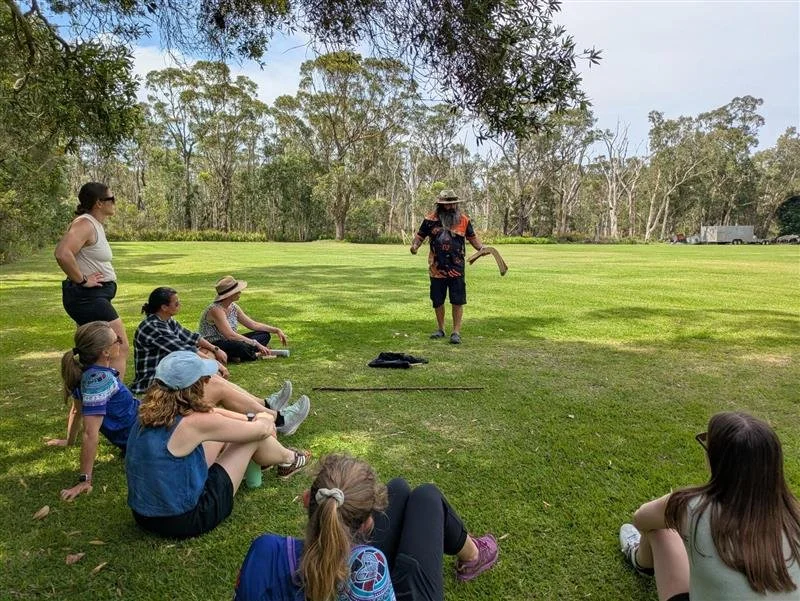 Group of children and adults outdoors watching a person dressed in colorful clothing and wearing a hat, performing or speaking in front of a grassy field with trees in the background.