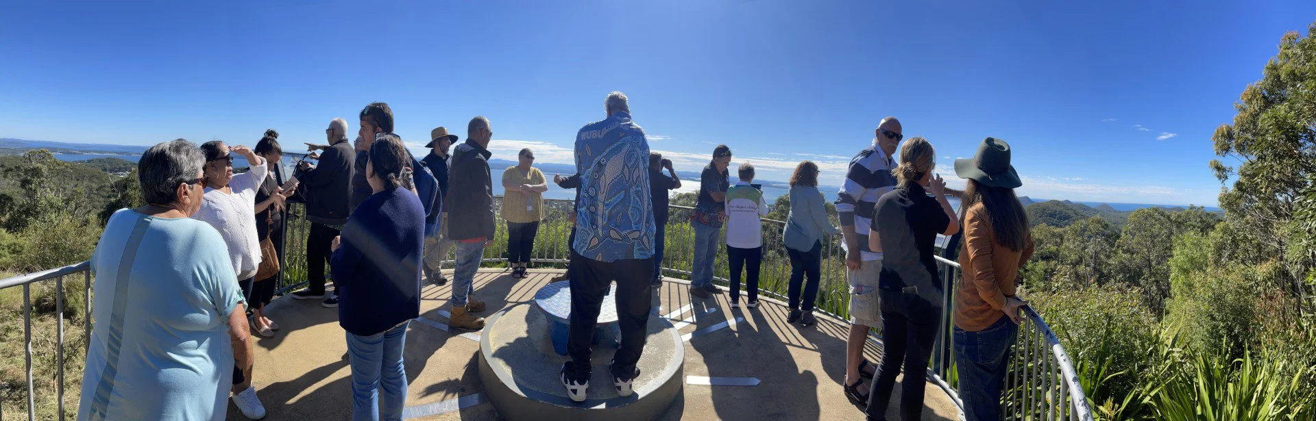 Group of people on a viewing platform overlooking a lush green landscape, some taking photos or enjoying the view, under a clear blue sky.