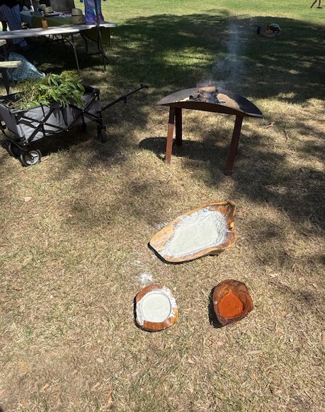 Outdoor scene with a small fire grill, ceramic dishes with white powder, and a cart with plants in a park.