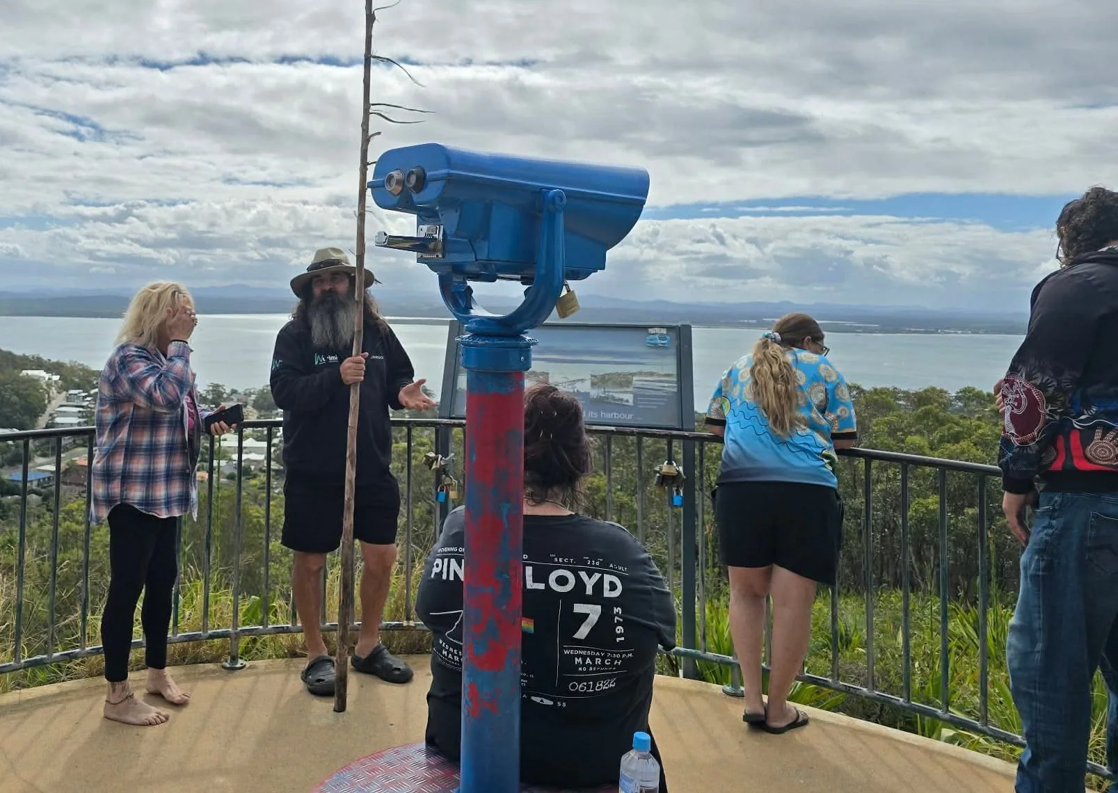 Group of people on a viewing platform overlooking a body of water with hills in the distance. Some are looking at a large coin-operated telescope, while others are talking or taking photos. The sky is partly cloudy.