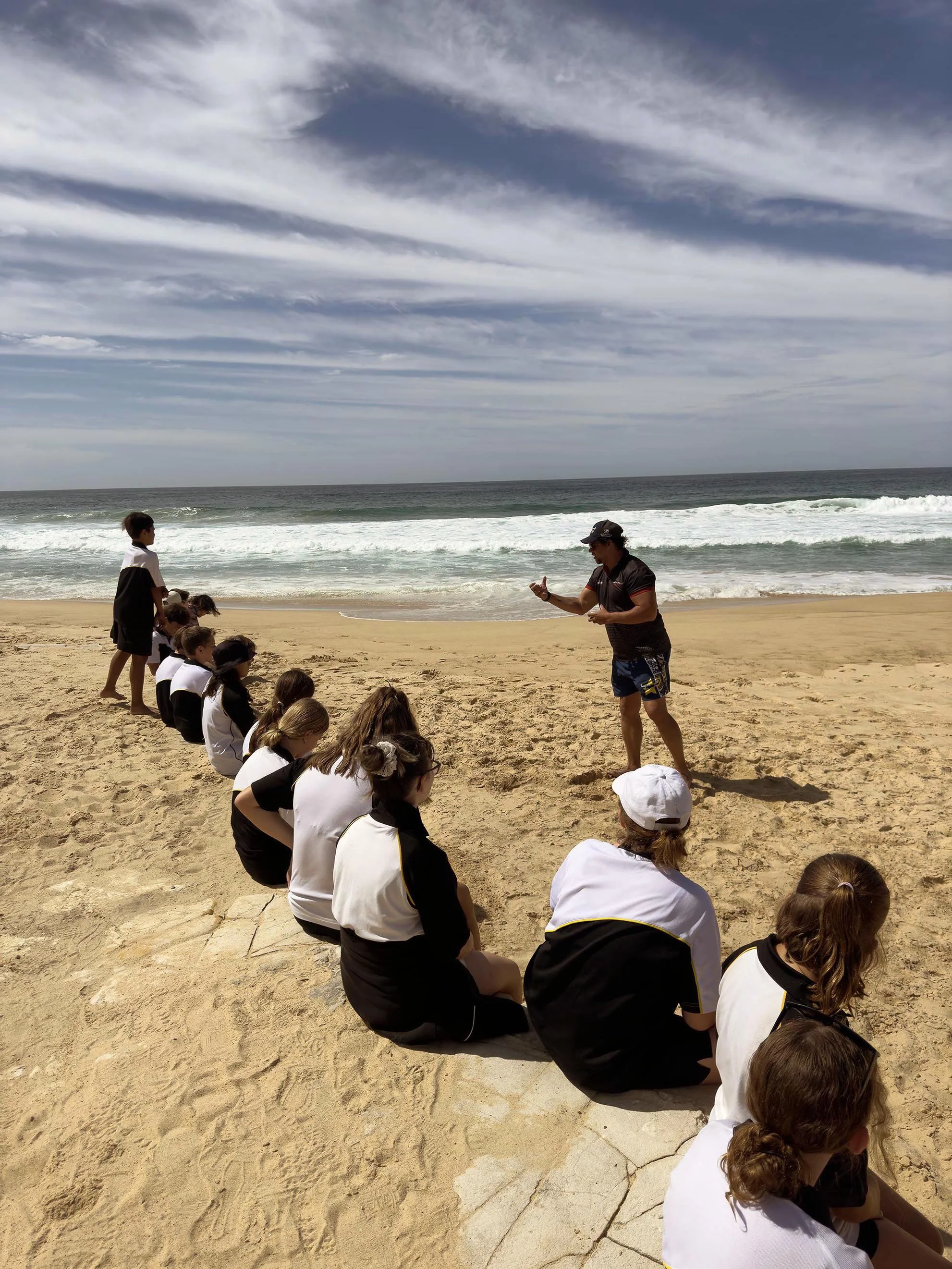 A group of children sitting on the sand facing the ocean, with an adult standing and talking in front of them at the beach under a partly cloudy sky.