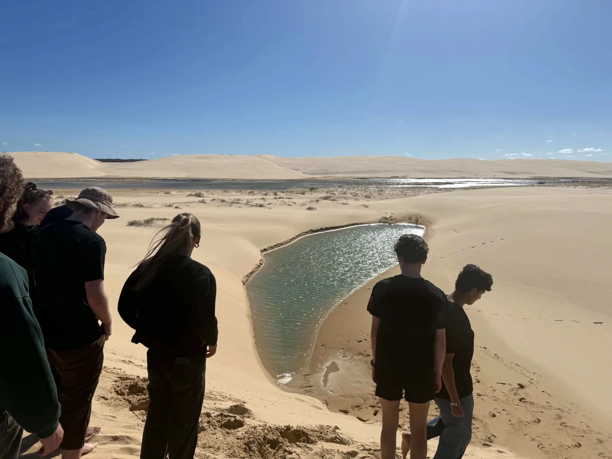 A group of people are standing on sandy dunes near a small body of water, under a clear blue sky.