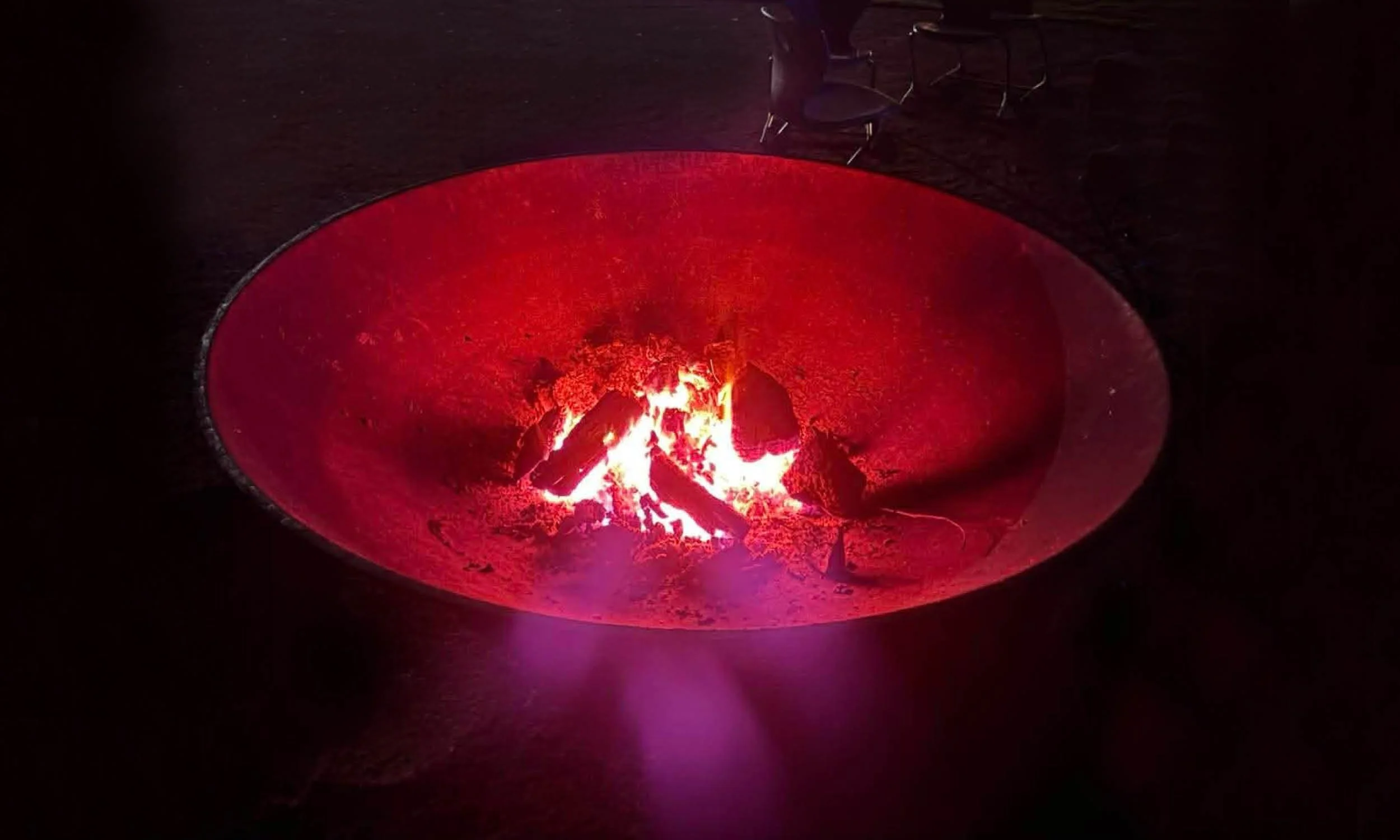 A fire burning inside a circular metal fire pit, with visible flames and logs, illuminated by red and orange glow in a dark surrounding.
