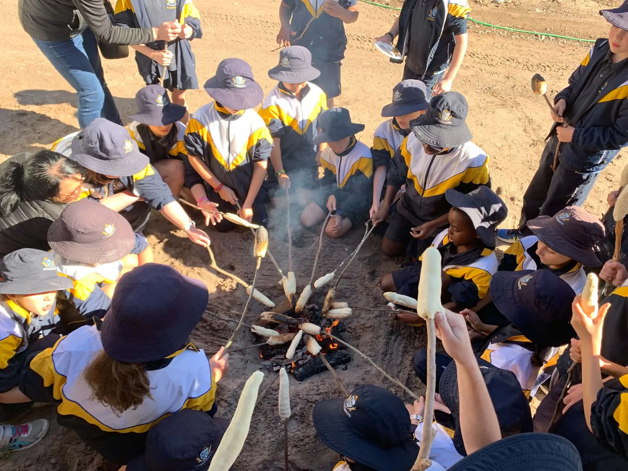 Group of children and adults gathered around bonfire, roasting marshmallows and hot dogs on sticks outdoors on sandy ground, likely at a camp.