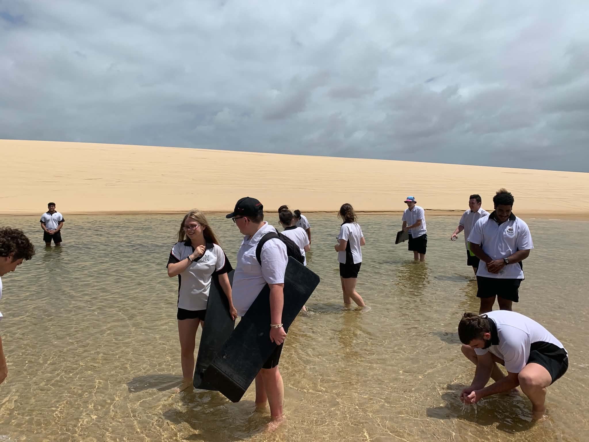 Group of people standing and walking in shallow water at the beach, with a large sand dune and cloudy sky in the background.